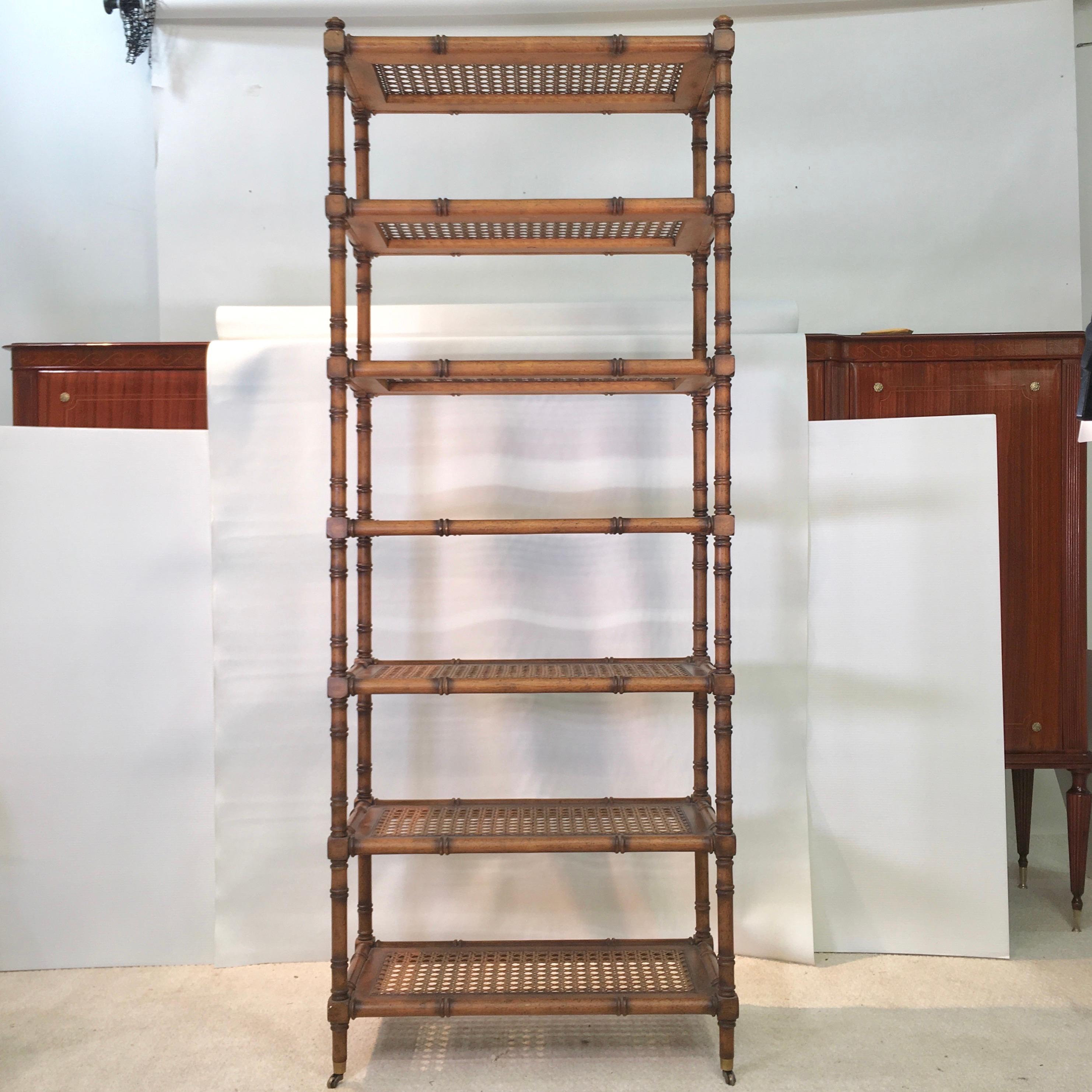 Hollywood Regency cum George IV-style étagère in faux bamboo wood in a provincial pecan finish and having seven caned open shelves, raised on four legs with brass casters. Finished on all sides so as to float freestanding within a room.