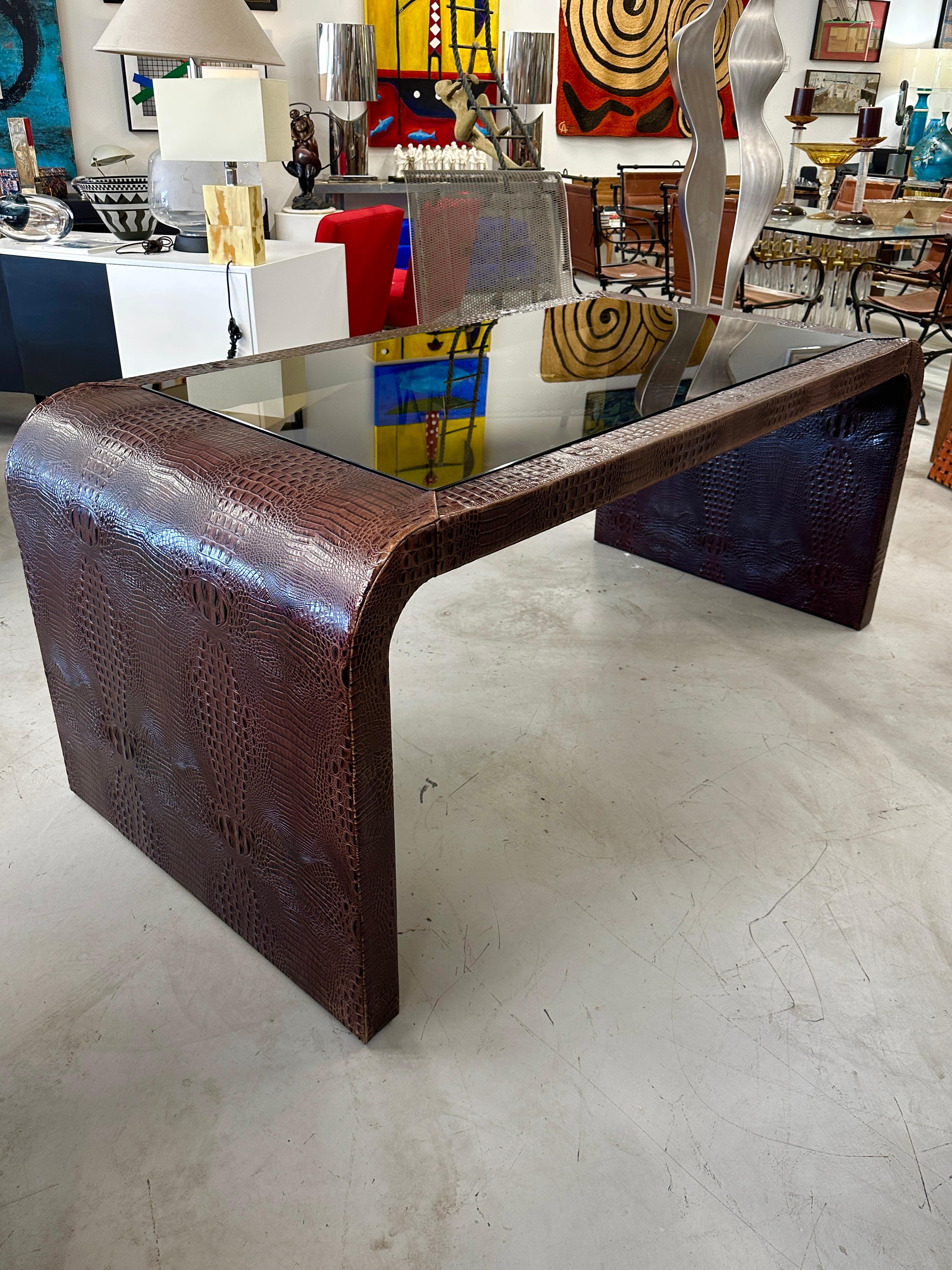 A stunning faux crocodile clad desk with a bronze glass inset. The leather is beautifully hand sewn on the desk and features striking graining. The clearance of the desk is 27.5 inches. It is pictured in our gallery with a Herman Miller Eames chair