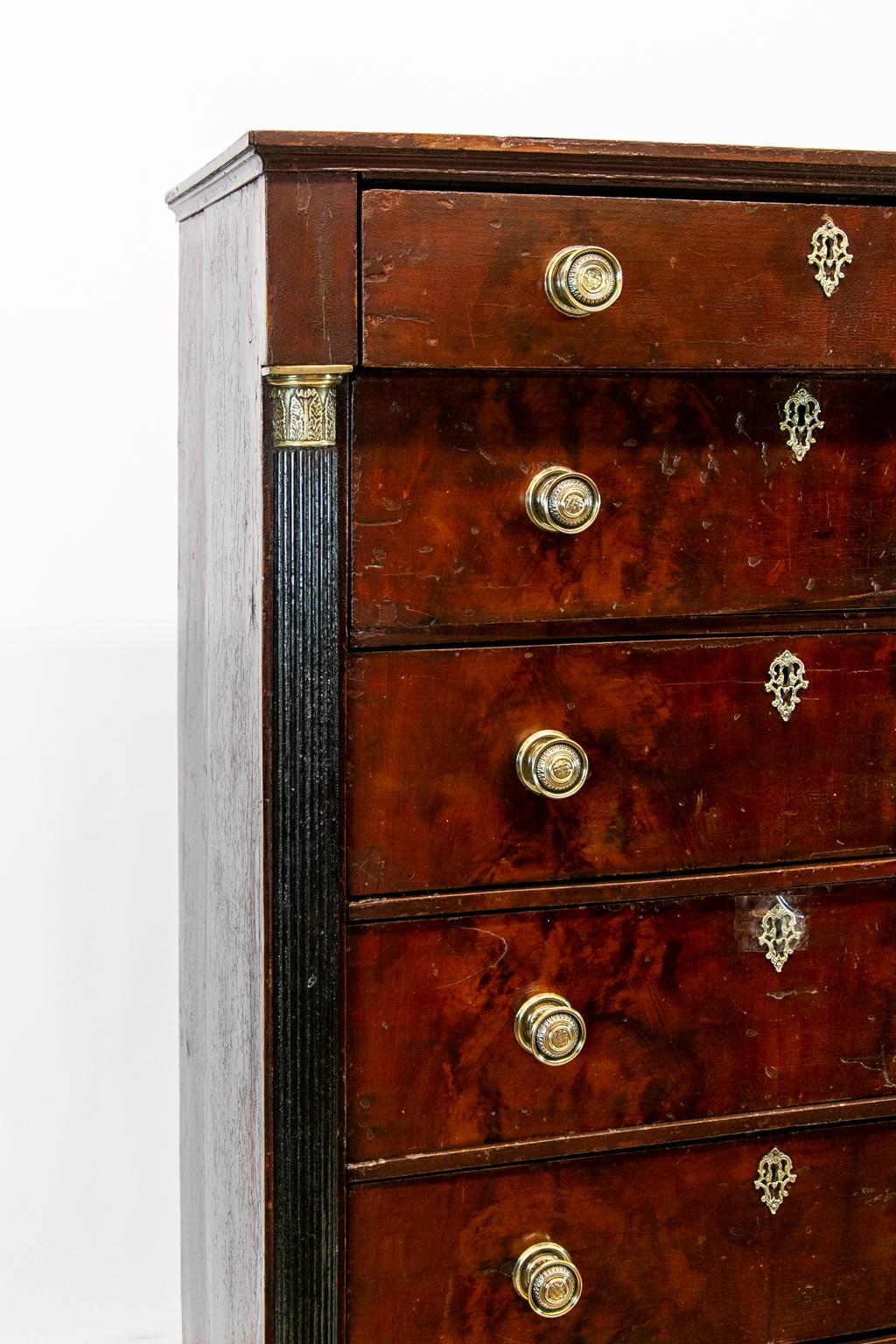 Faux painted six-drawer chest is painted to simulate croch mahogany. The stiles have fluted palisters with brass capitals and bases terminating in tapered feet. The brass knobs and escutcheons are correct for the period but are not original.