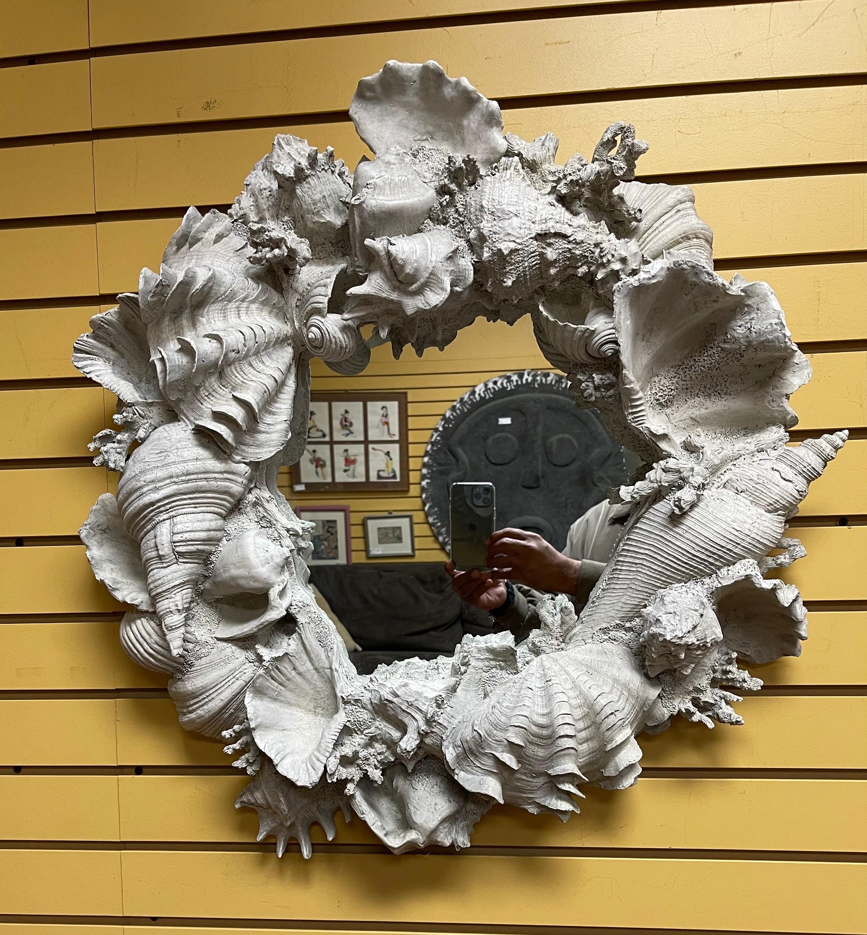 A very cool faux seashell encrusted mirror in molded fiberglass, circa 2000s. The mirror is decorated throughout with shells of various sizes and designs; it has a concrete look and color to it. The piece measures: 28