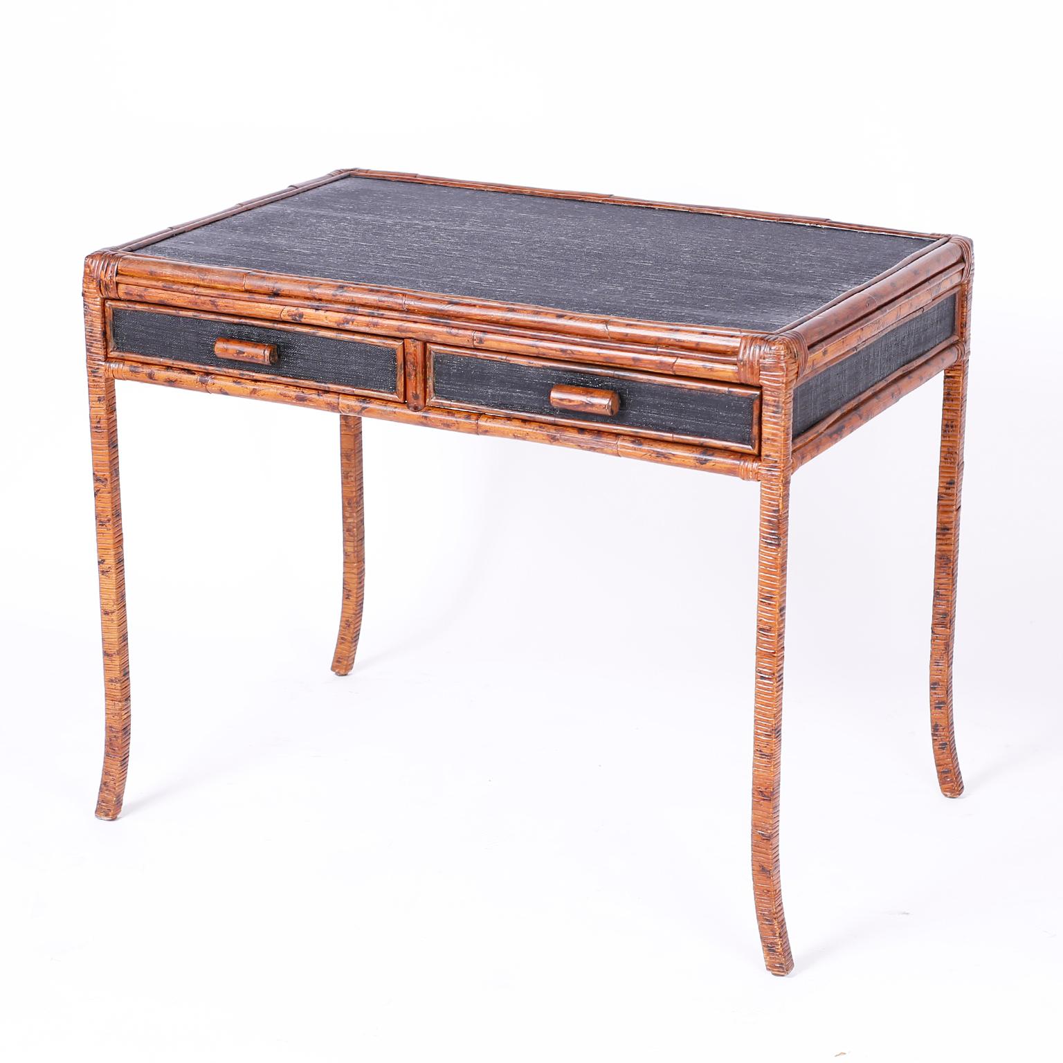 Midcentury British colonial two-drawer writing desk with black painted grass cloth panel on the top, front, sides, and back. The frame is faux bamboo with a tortoise finish and reed wrapped elegant metal splayed legs.