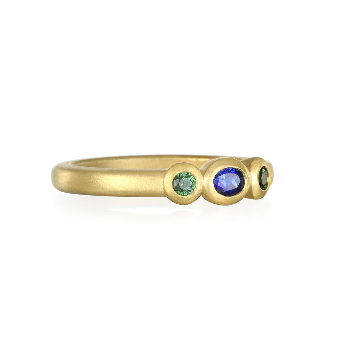 A vibrant oval shaped blue sapphire stone flanked by sparkling green sapphires. This three stone 18 karat gold Faye Kim ring is beautifully crafted, bezel set and matte-finished.  Wear alone or stacked with other rings to elevate your individual