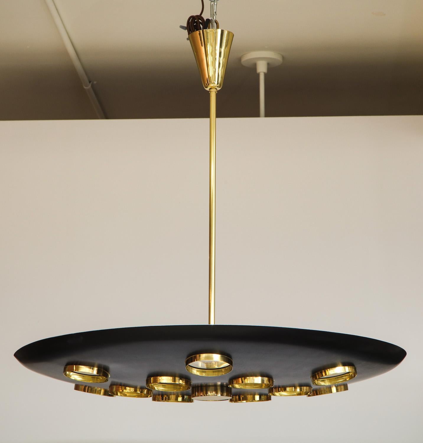 Black-painted aluminum saucer form, with circular cut-outs. Each cut-out is fitted with a brass framed glass lens, creating a subtle abstract effect. 6 standard E26 sockets and polished brass mounts. The rod can be cut down and this light can be