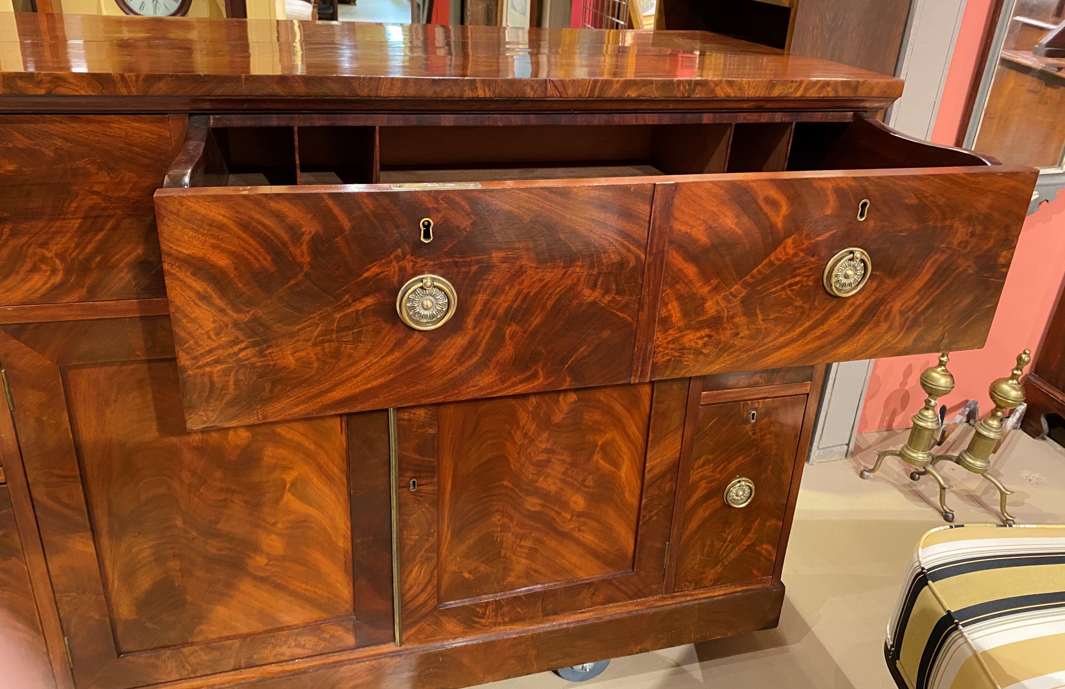 A fine example of a figured mahogany veneered sideboard or server with fitted hidden butler’s desk with drop down leather writing surface and compartmentalized interior, surmounting two paneled doors, opening to interior shelved storage