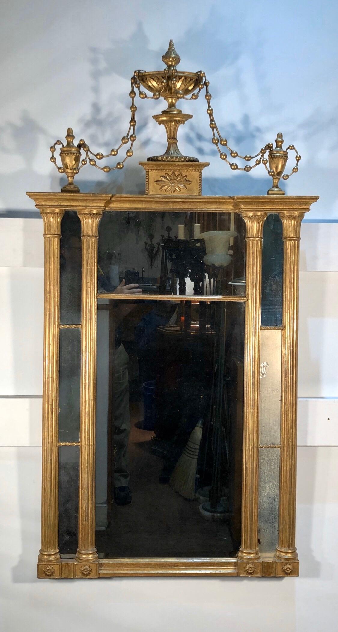 This Classic Greek and Egyptian motif mirror has vertical pillars flanking mirror panels framing the central split looking glass. The cornice of the mirror has a Classic Adams Urn on plinth with a rosette flanked by matching Adams Urns draped in