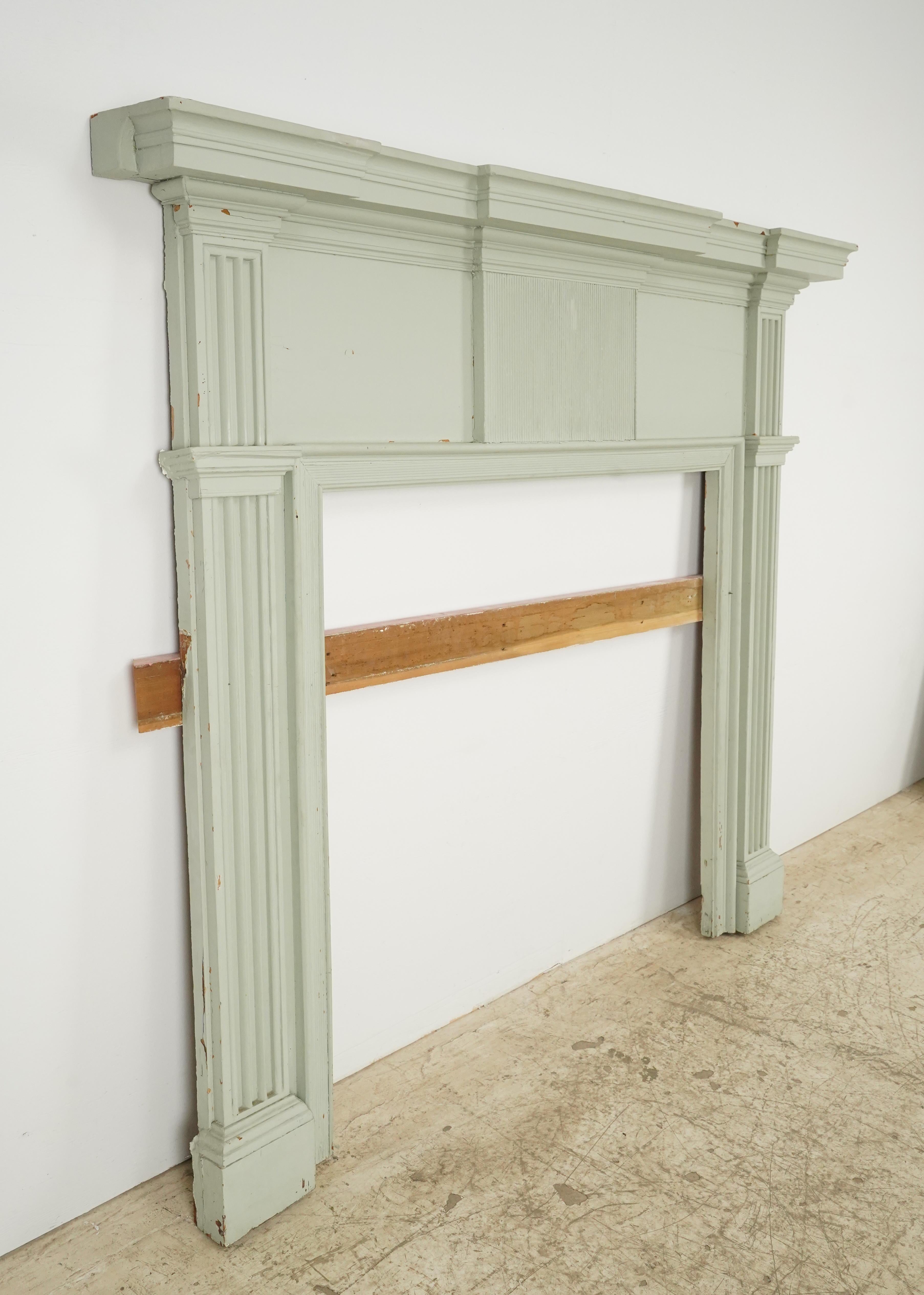 Cheminée de style Feder en bois peint de couleur vert-gris. Elle présente des pilastres proéminents de part et d'autre, qui soutiennent une importante tablette de cheminée et un frise. Les pilastres ont des cannelures verticales et le panneau de la