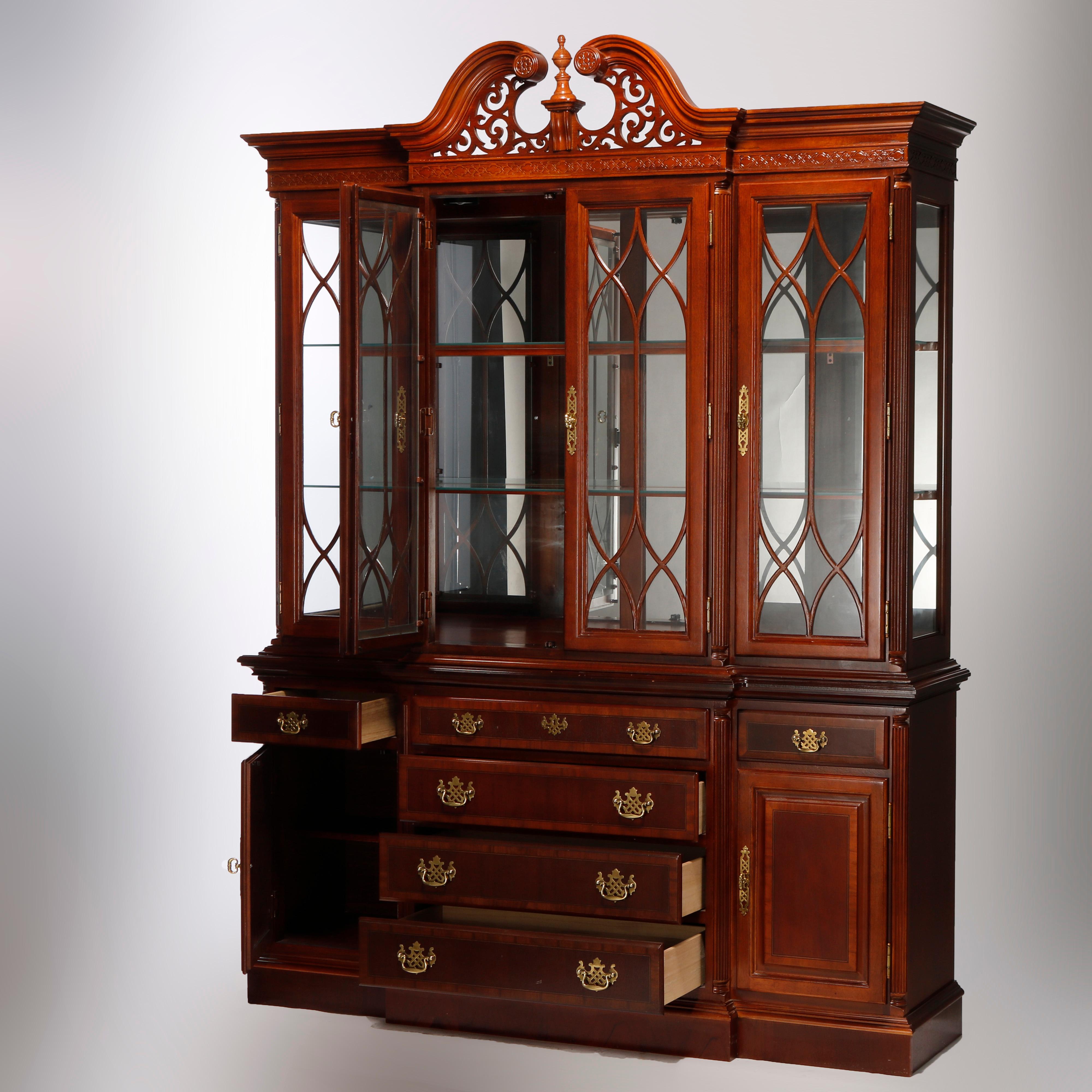A Federal style breakfront cabinet in the manner of Baker Furniture offers broken arch crest with rosettes, foliate filigree and central turned finial over upper having four glass doors, each with stylized mullion decoration and opening to shelved,