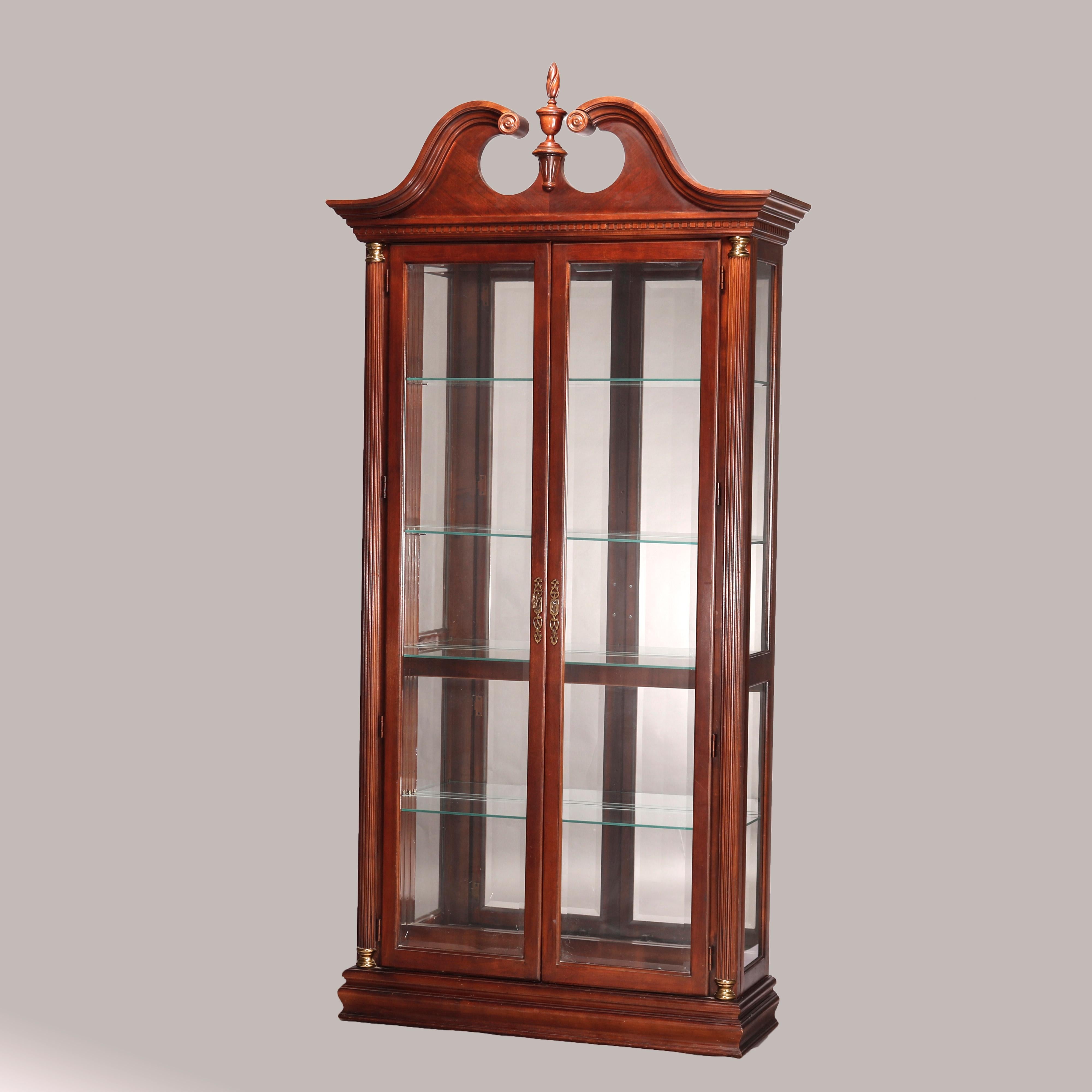 A Federal style display case by Pulaski offers mahogany construction with broken arch crest having central carved finial over case having double glass doors opening to shelved and mirrored interior, flanking reeded Greco Doric style columns with