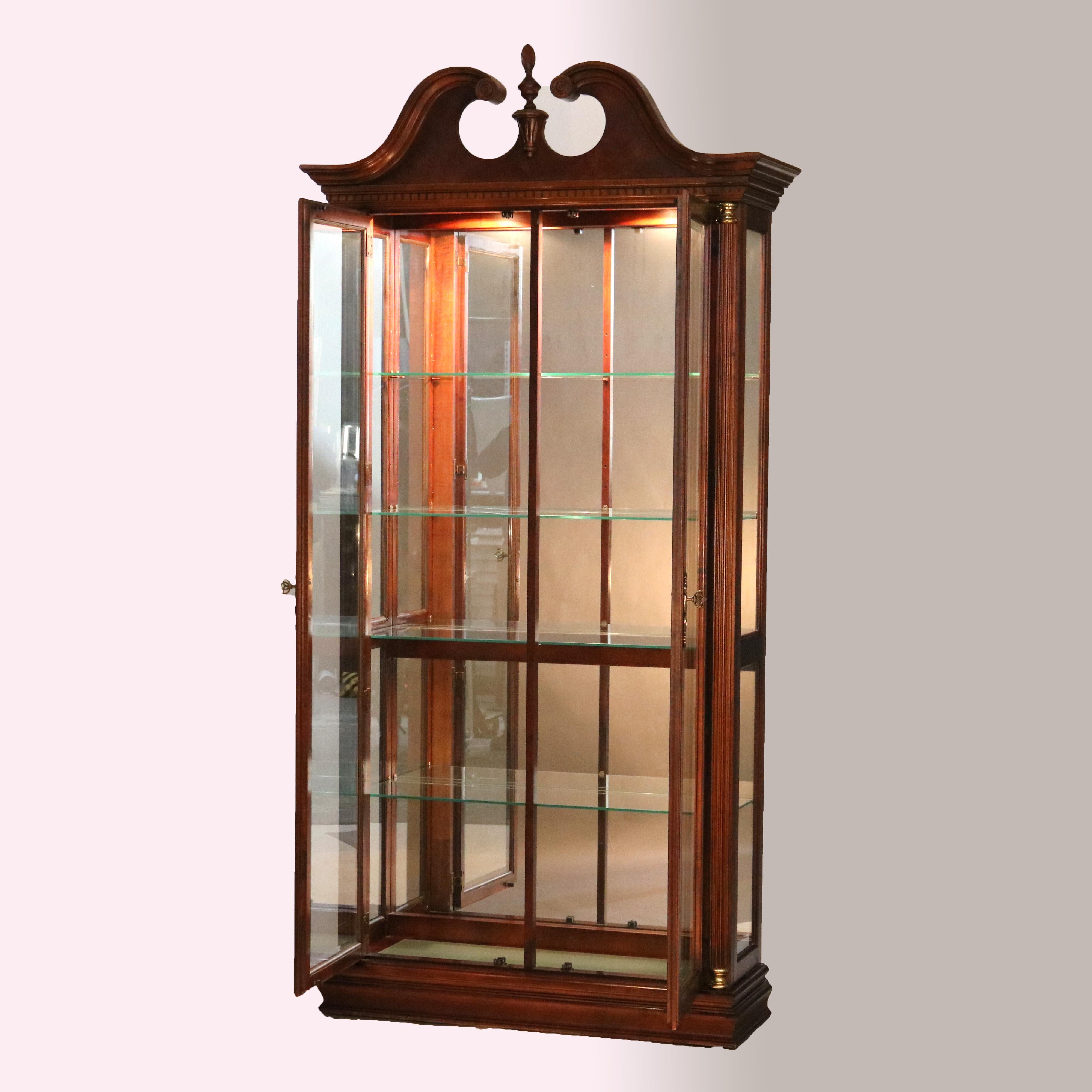 A Federal style display case by Pulaski offers mahogany construction with broken arch crest having central carved finial over case having double glass doors opening to shelved and mirrored interior, flanking reeded Greco Doric style columns with