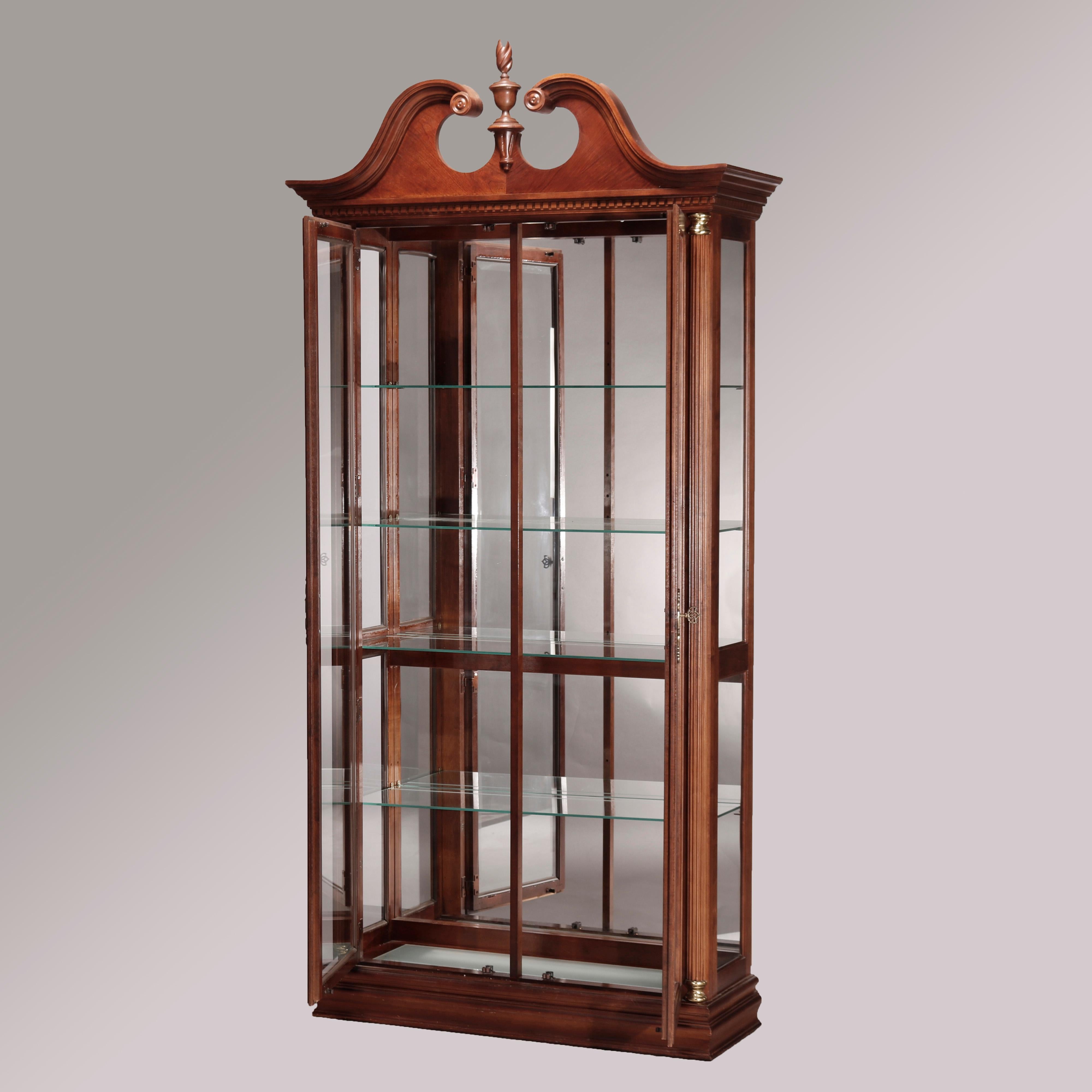 A Federal style display case by Pulaski offers mahogany construction with broken arch crest having central carved finial over case having double glass doors opening to shelved and mirrored interior, flanking reeded Greco Doric style columns with