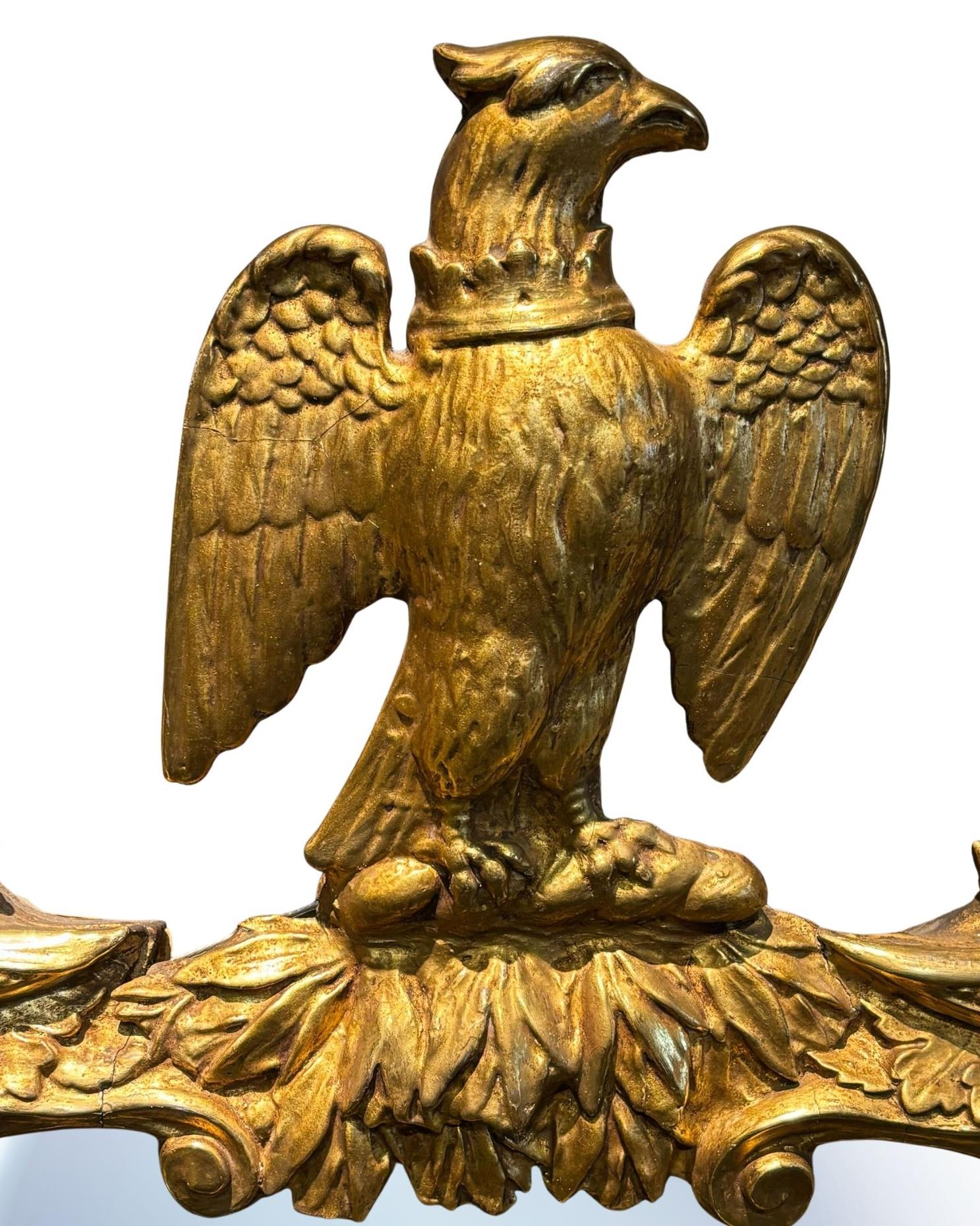 Se presenta un espejo de estilo federal con un impresionante remate de águila. El marco del espejo está ornamentado y pintado de dorado. En la parte superior hay una gloriosa águila de alas extendidas posada sobre un manojo de hojas. Los laterales