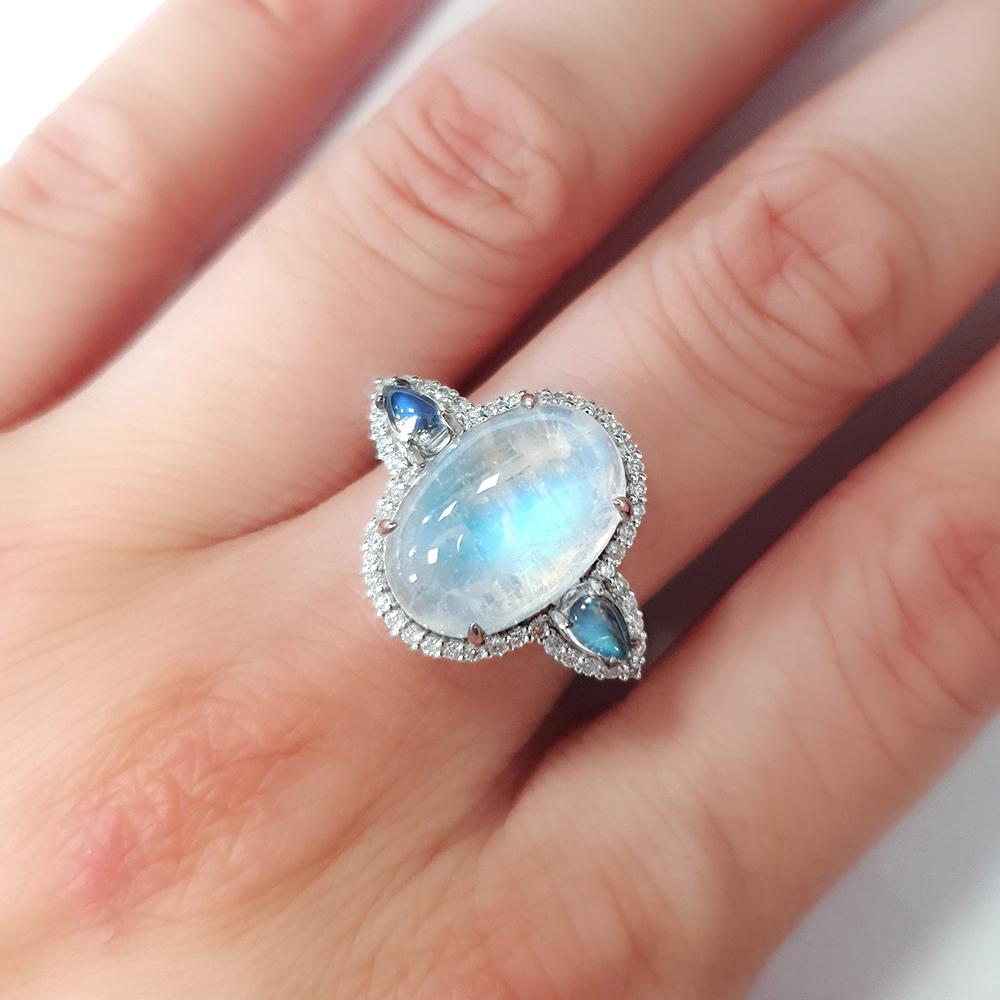 Crafted in luxurious platinum, this iridescent ring features a captivating 7.88ct arrangement of moonstones, with a luminous central cabochon flanked by two pear-shaped accents that shimmer with ethereal blue flashes. A halo of 0.276ct sparkling