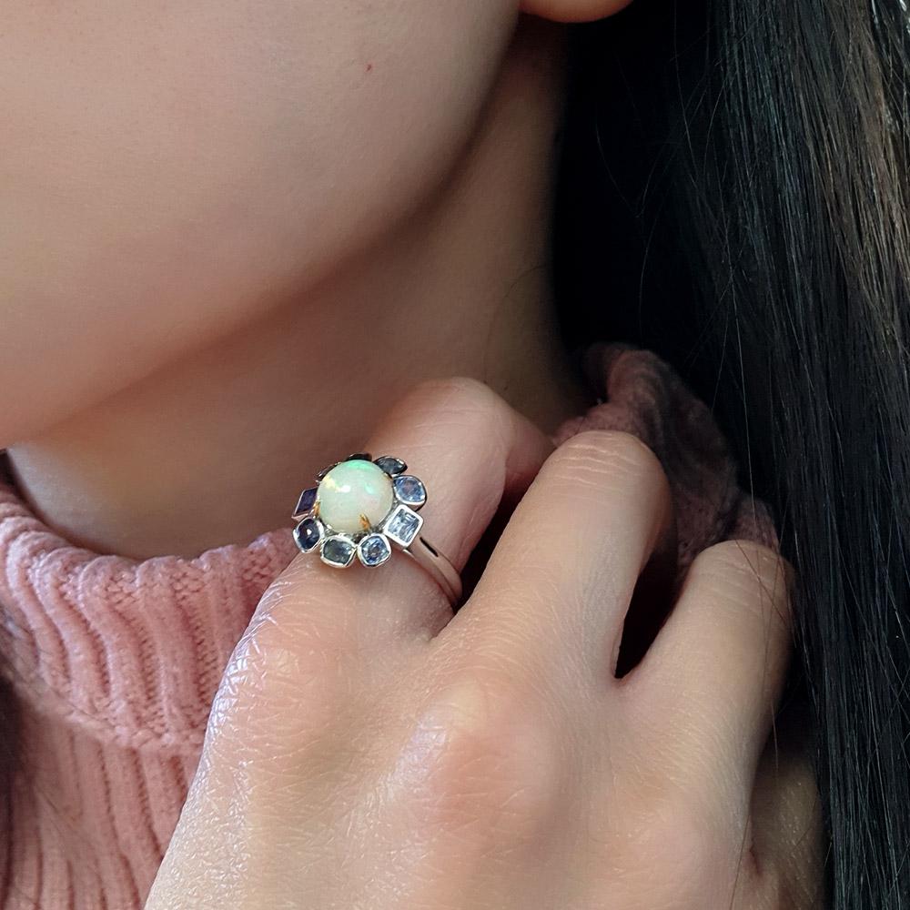 Indulge in the opulent allure of this exquisite ring, boasting a central 1.33ct opal, encircled by a beautiful assortment of sapphires, totalling 1.4ct in weight, meticulously crafted in an 18ct white gold setting with delicately rose gold-plated