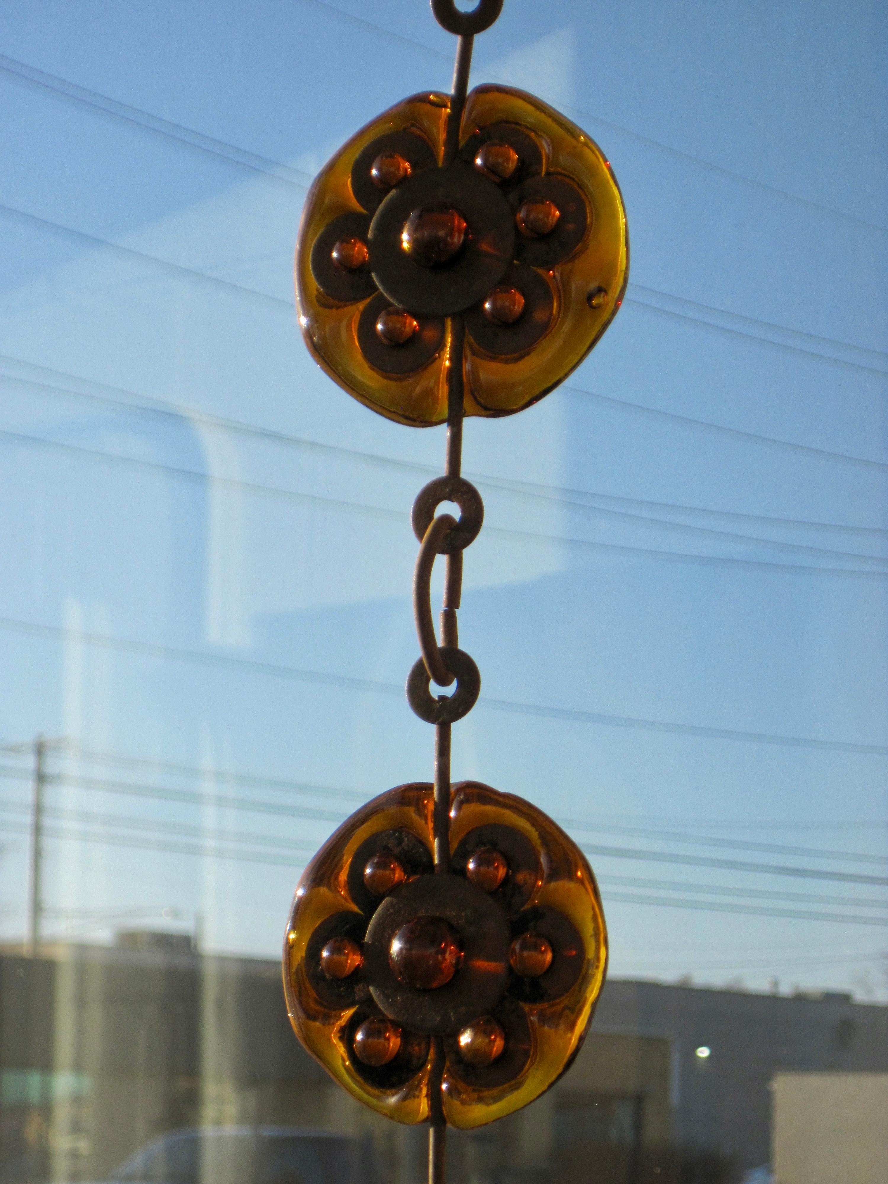A Felipe Delfinger sun catcher made of amber art glass sculpted and protruding through iron circles creating daisy-like round flowers. Eleven flowers attached by iron rings hang down 76 inches long, catching the sun beautifully and the patinated