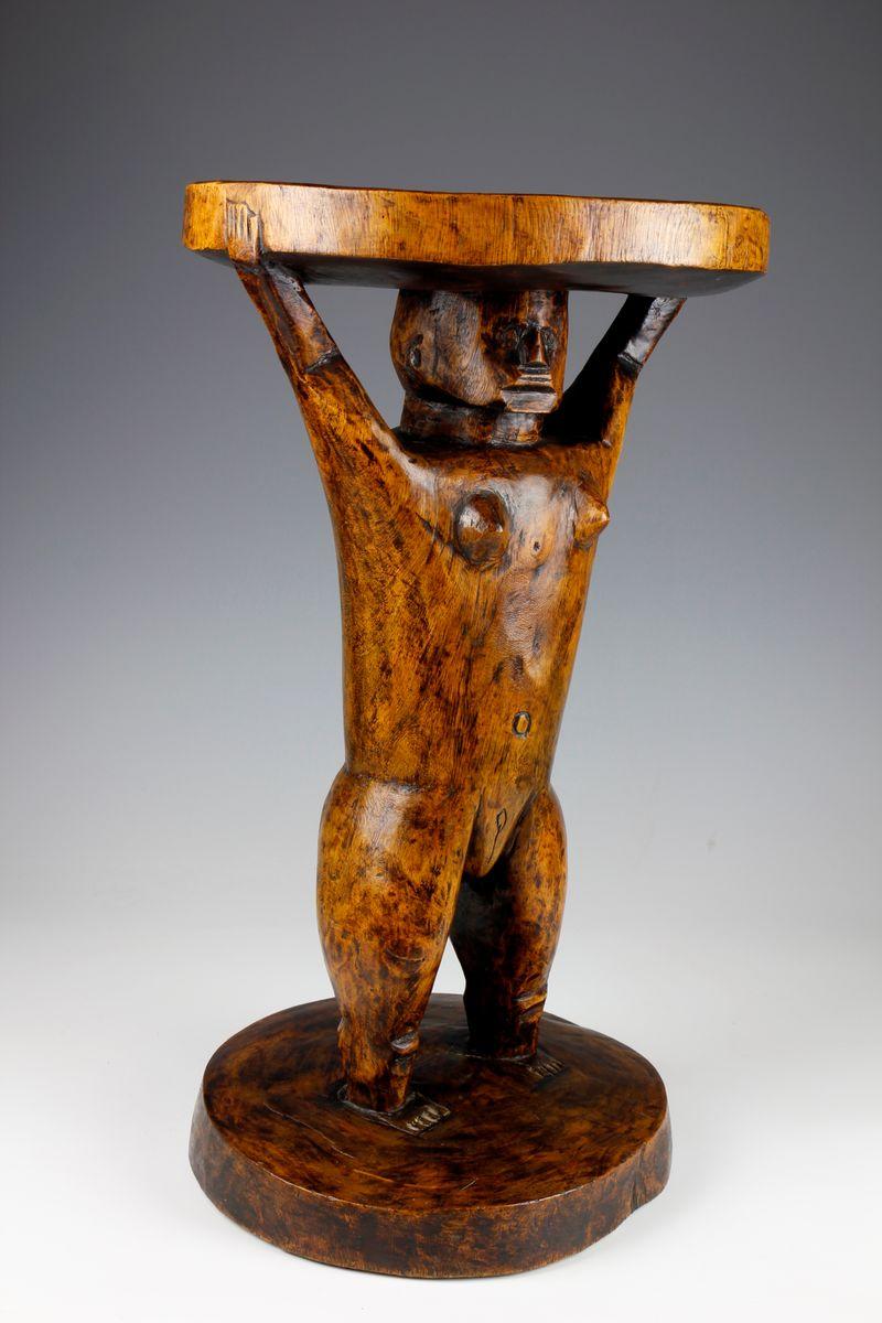 Auf der flachen, runden Sitzfläche dieses Hockers aus der Shona-Kultur in Simbabwe aus dem frühen zwanzigsten Jahrhundert steht eine figurative Skulptur einer kräftigen Frau. Die Figur ist mit erhobenen Armen dargestellt, wobei sie den Sitz über