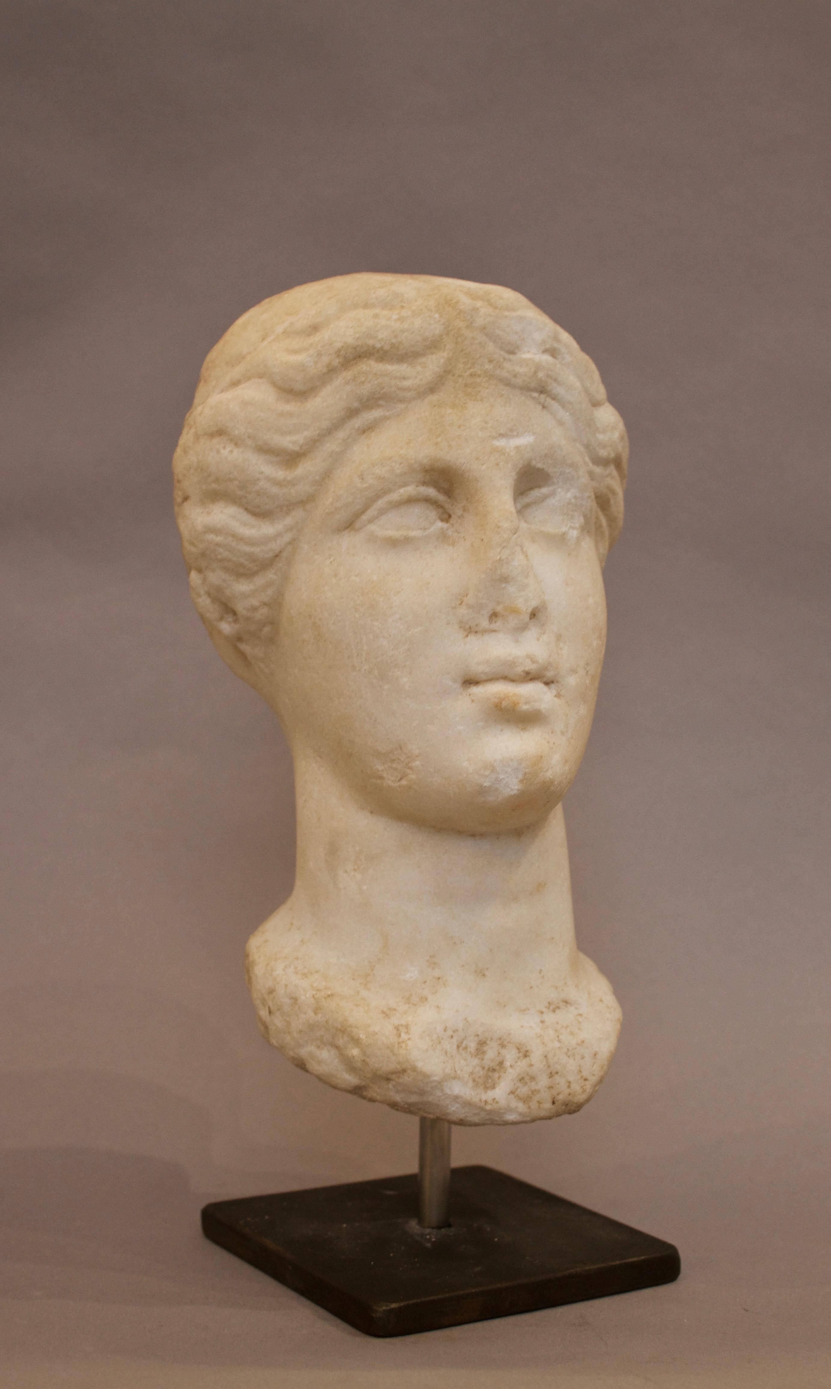An elegant oval marble feminine head dating from the Hellenistic period, which features a neat and almost symmetrical parted hairstyle that covers the entirety of the ears. The woman seems to gaze slightly upwards and features a closed mouth. The