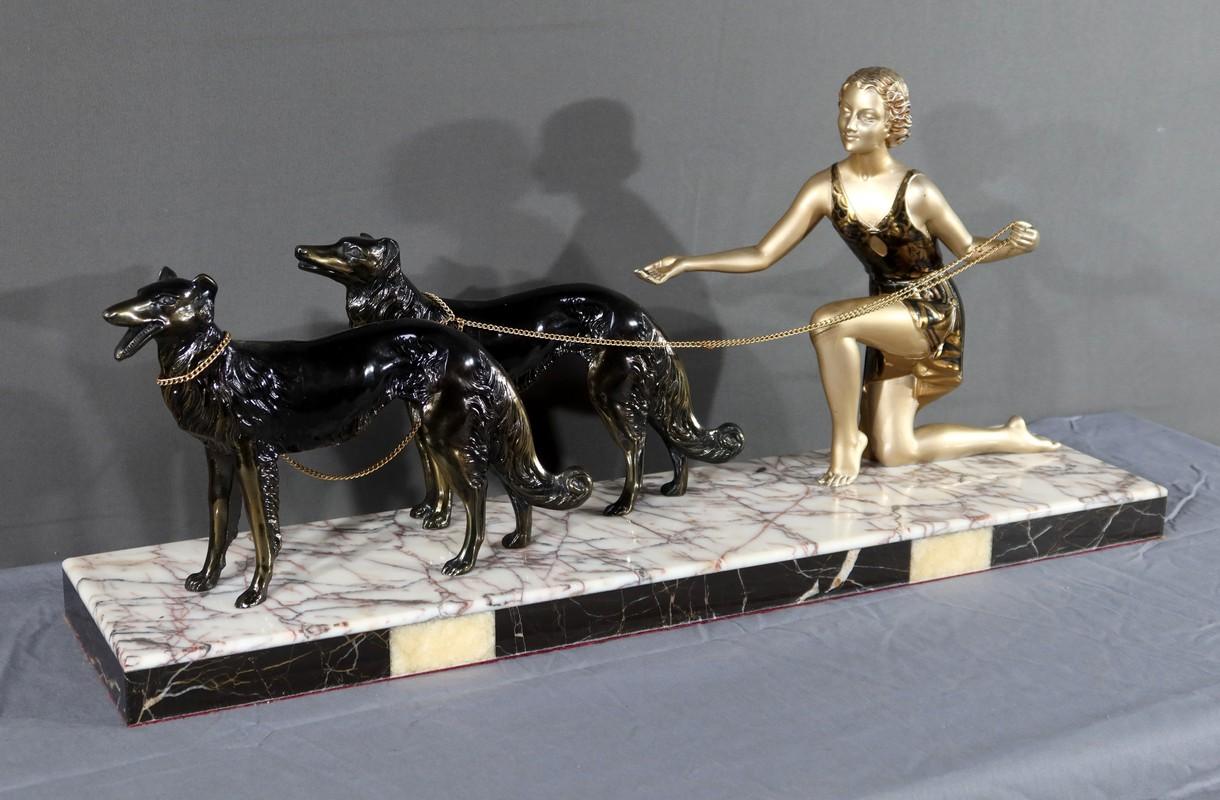Dieses Werk zeigt eine Frau in Begleitung von zwei Windhunden. Es ist aus Metall mit schwarzer und goldener Patina gefertigt.
Die junge Frau geht mit ihren Hunden an der Leine spazieren, wie die goldfarbene Kette andeutet, die sie miteinander