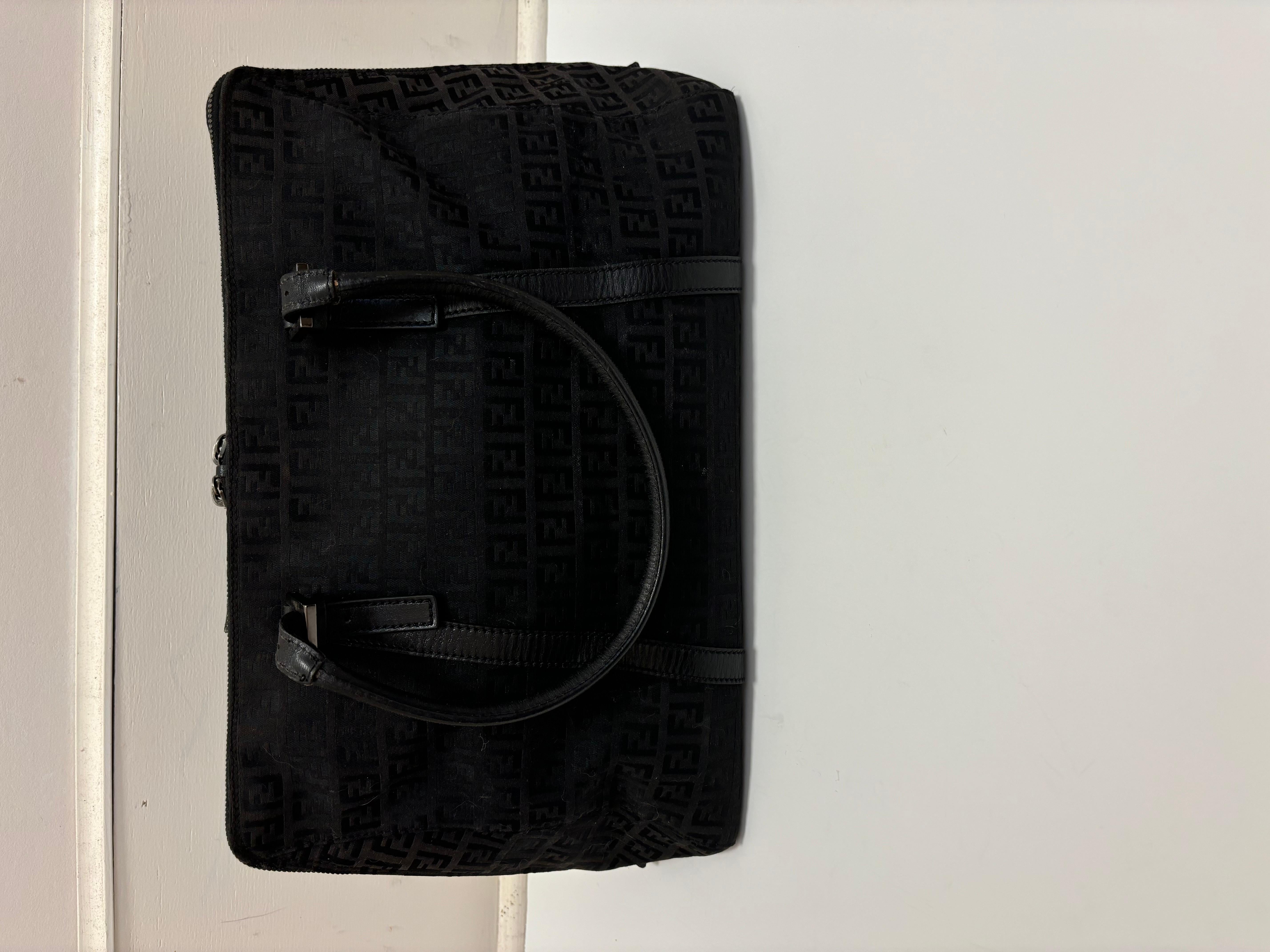 Ce top handle/tote est réalisé en toile monogramme Zucca noire avec des poignées rondes, une bordure et un côté en cuir. Une poche intérieure zippée est doublée de tissu noir Fendi. Le matériel argenté gravé par Fendi comprend les tirettes de la