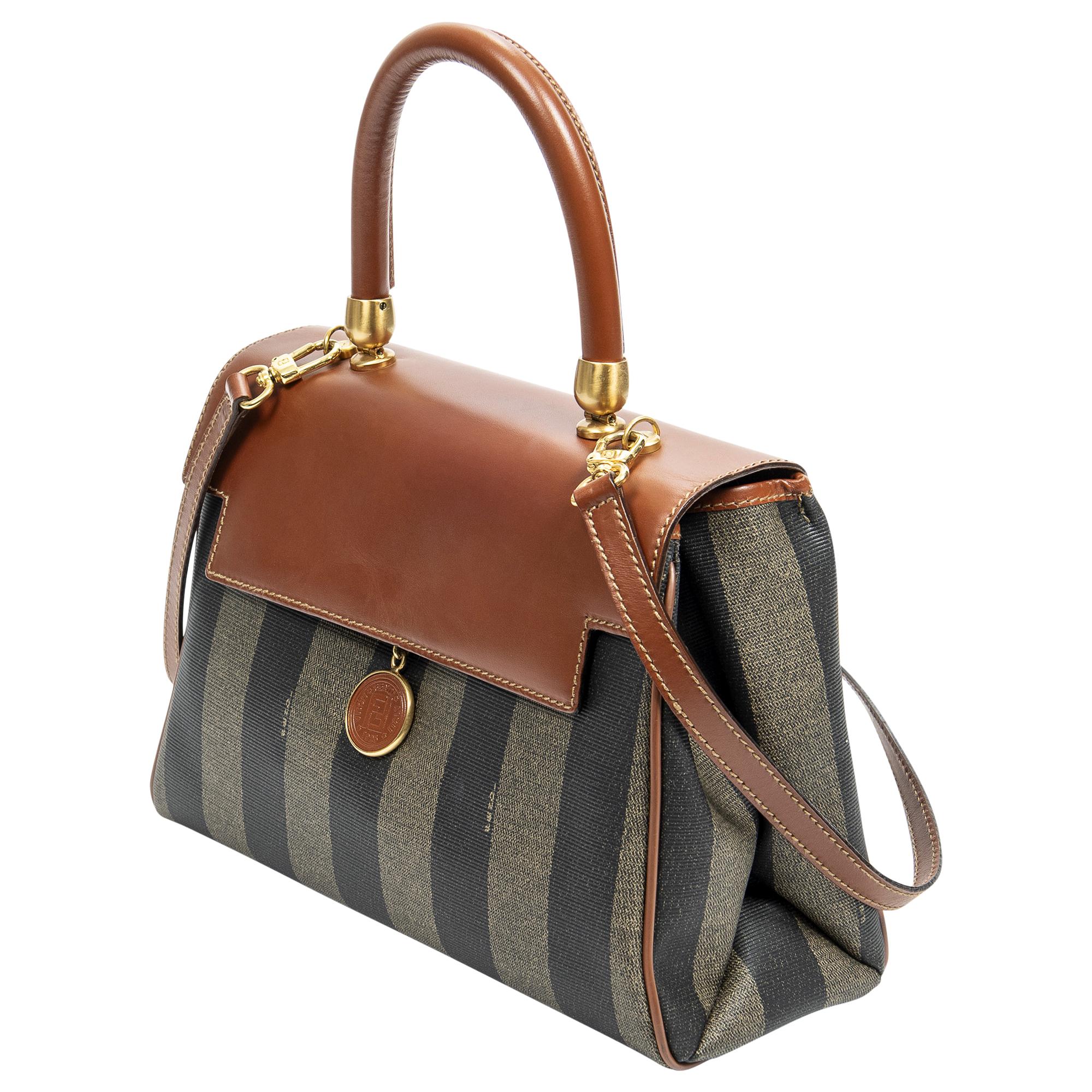 Introducing the Fendi Brown Vintage Pequin Top Handle Bag, a timeless piece for the discerning fashion aficionado. Crafted in durable brown coated canvas, this bag boasts an elegant gold clasp closure that adds a touch of luxury. The sturdy top
