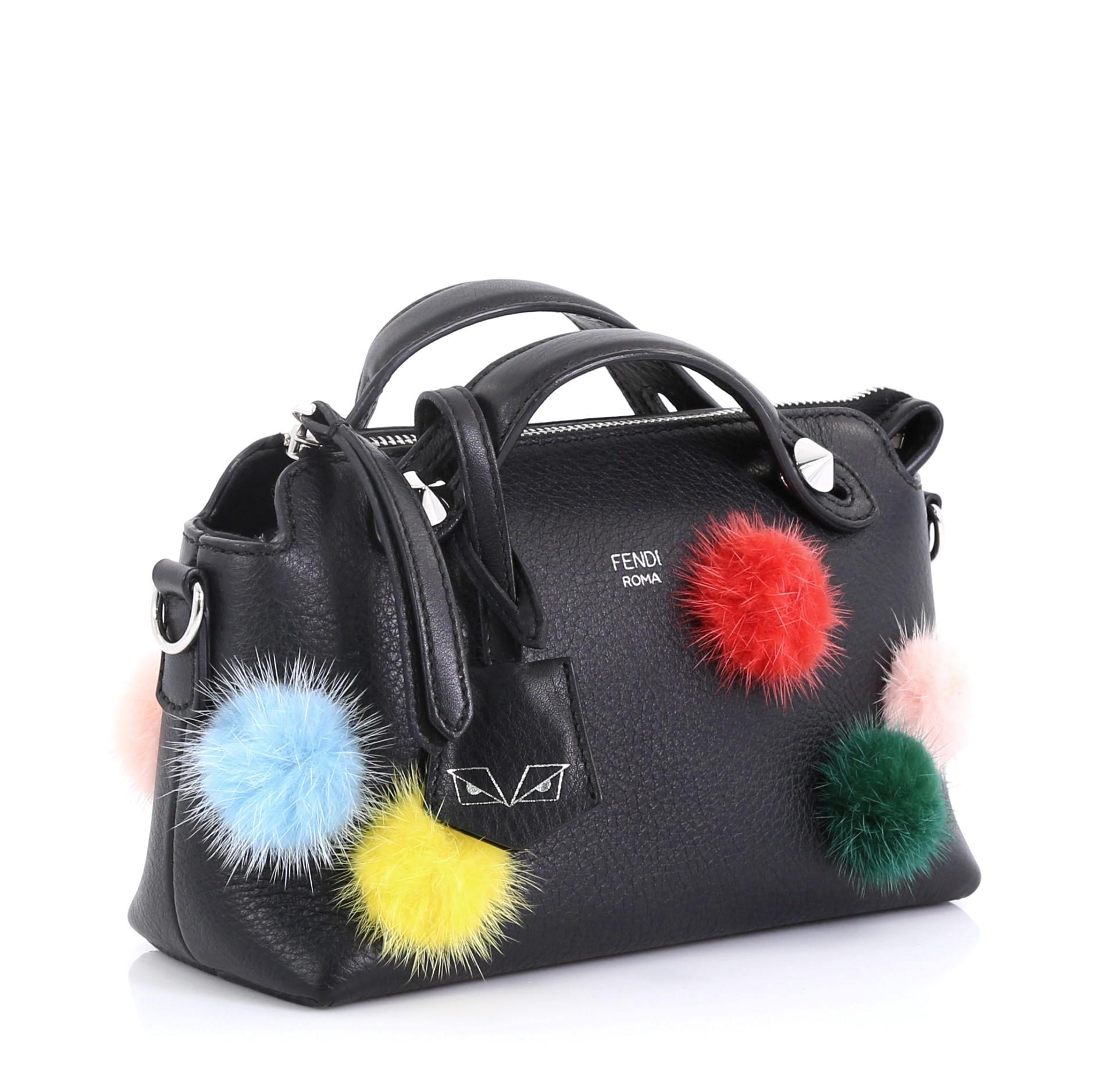 This Fendi By The Way Satchel Pom Pom Leather Mini, crafted from black leather, features dual flat top handles with cone studs, adjustable shoulder strap, dyed Denmark mink fur pompoms, and silver-tone hardware. Its top zip closure opens to a black