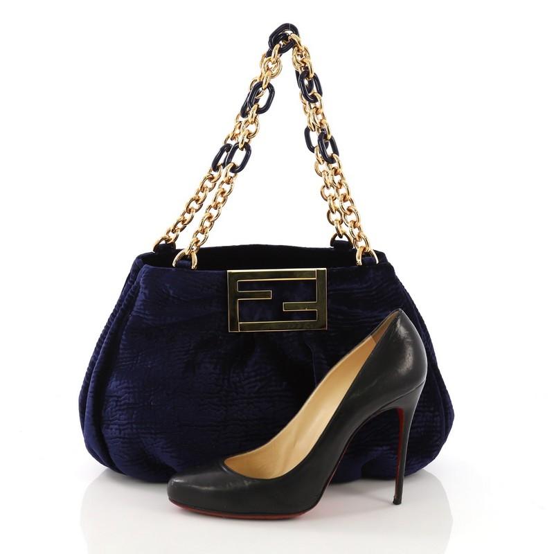 This Fendi Mia Tote Velvet Small, crafted from blue velvet, features woven-in leather chain handles, giant Fendi logo, and gold-tone hardware. Its wide top opening showcases a brown satin interior with middle zip compartment and two open