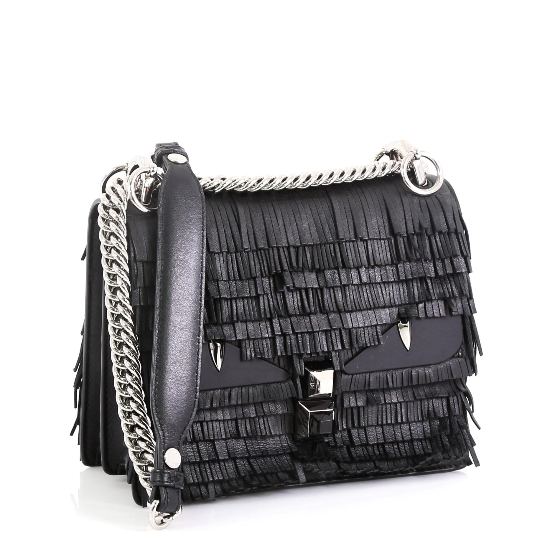 This Fendi Monster Kan I Bag Fringe Leather Small, crafted from black fringe leather, features chain link strap with leather pad, monster eye applique design, and silver-tone hardware. Its plexiglass twist-lock closure opens to a gray microfiber