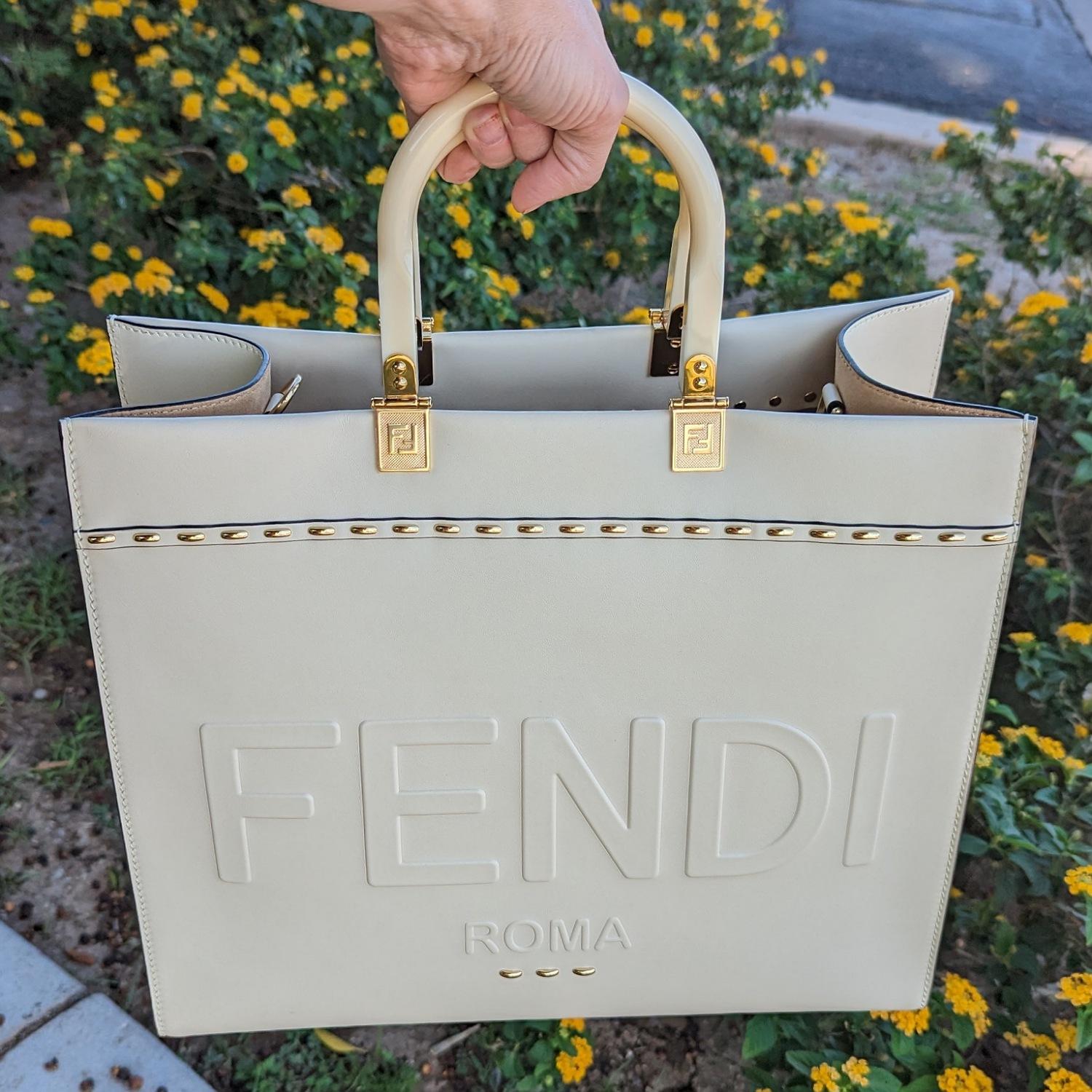Medium Sunshine Shopper bag made of creme leather with heat-stamped “FENDI ROMA” and stiff plexiglass handles. Edges in tone on tone leather with gold-finish metal hardware. Can be carried by hand or worn on the shoulder thanks to the two handles