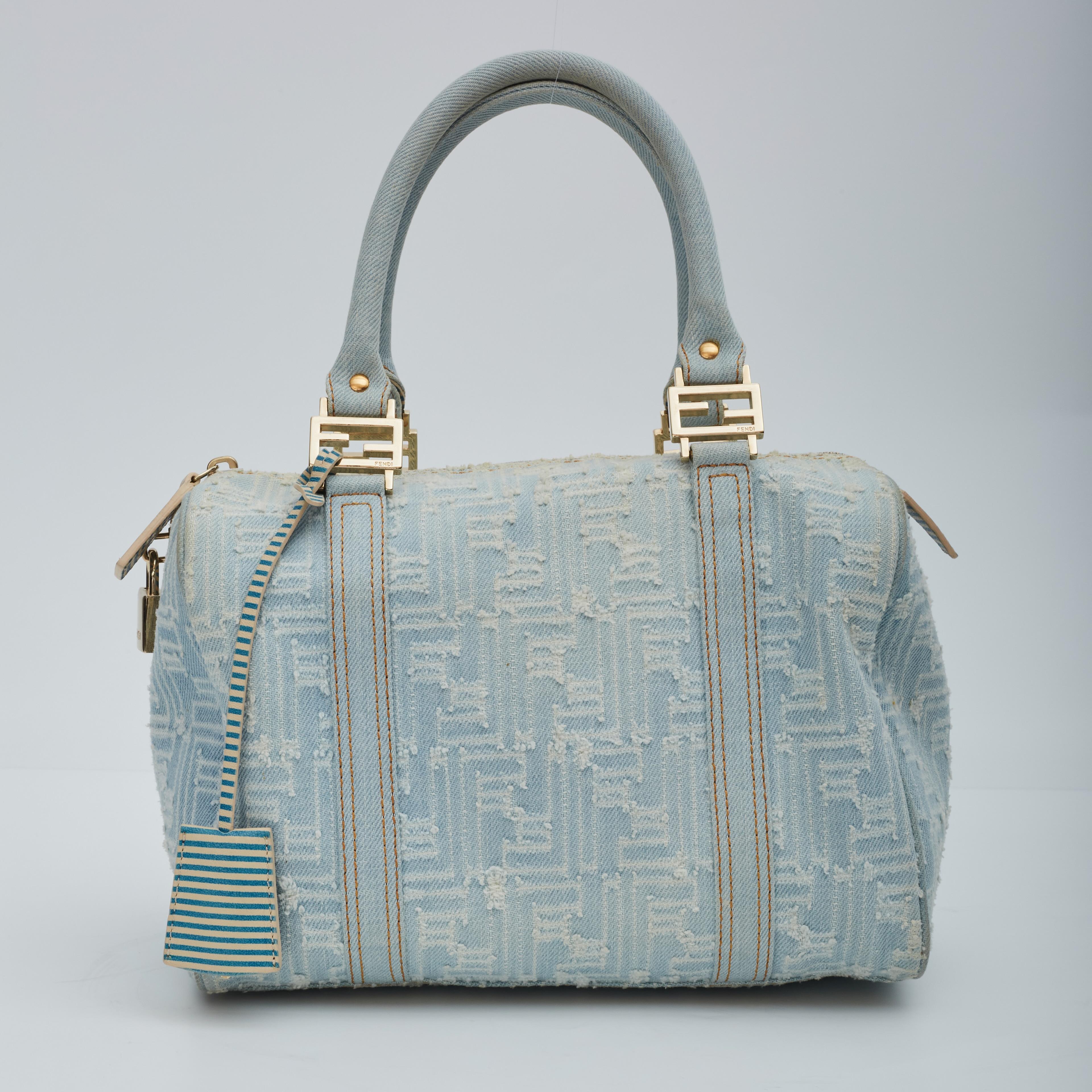 This Fendi bauletto Boston 25cm handbag is made with distressed blue denim with white stitched Fendi Zucca monogram pattern. Featuring gold-tone hardware, dual rolled top handles, logo buckle accents, cerulean and creme striped leather finishes,