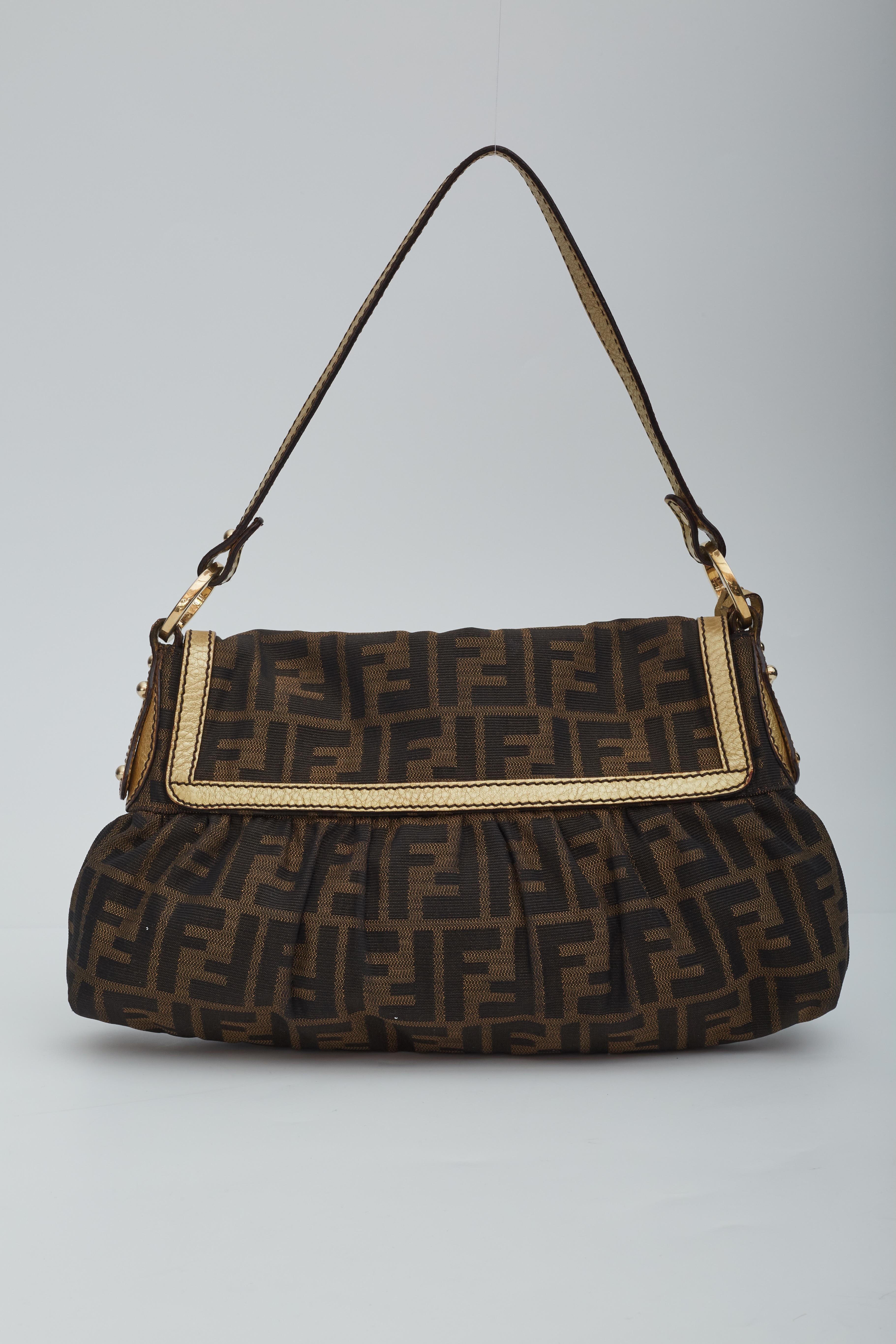 This bag is made with classic Fendi monogram Zucca print on brown canvas with gold leather finishes. The bag features a flat leather shoulder strap attached by gold tone hardware circular links, a hanging logo charm disk at front, a front flap, and