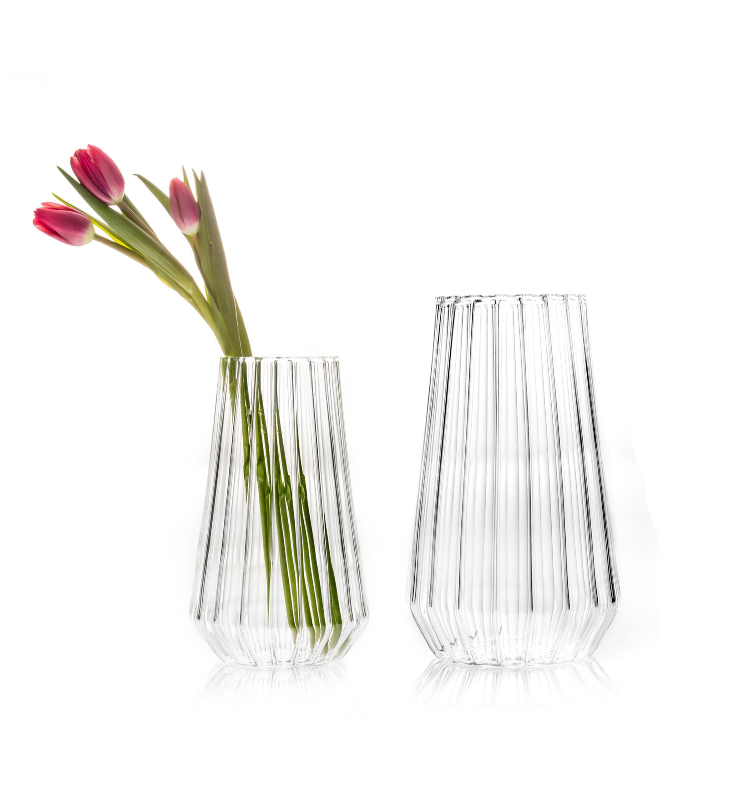 Stella Vasen Large & Medium - 2er-Set

Die reine Form, der linsenförmige Effekt des geriffelten Glases verdeckt die Stiele und bringt die Blütenköpfe zur Geltung. Die Facette in der Nähe des Bodens dient als Markierung für die Wasserlinie und