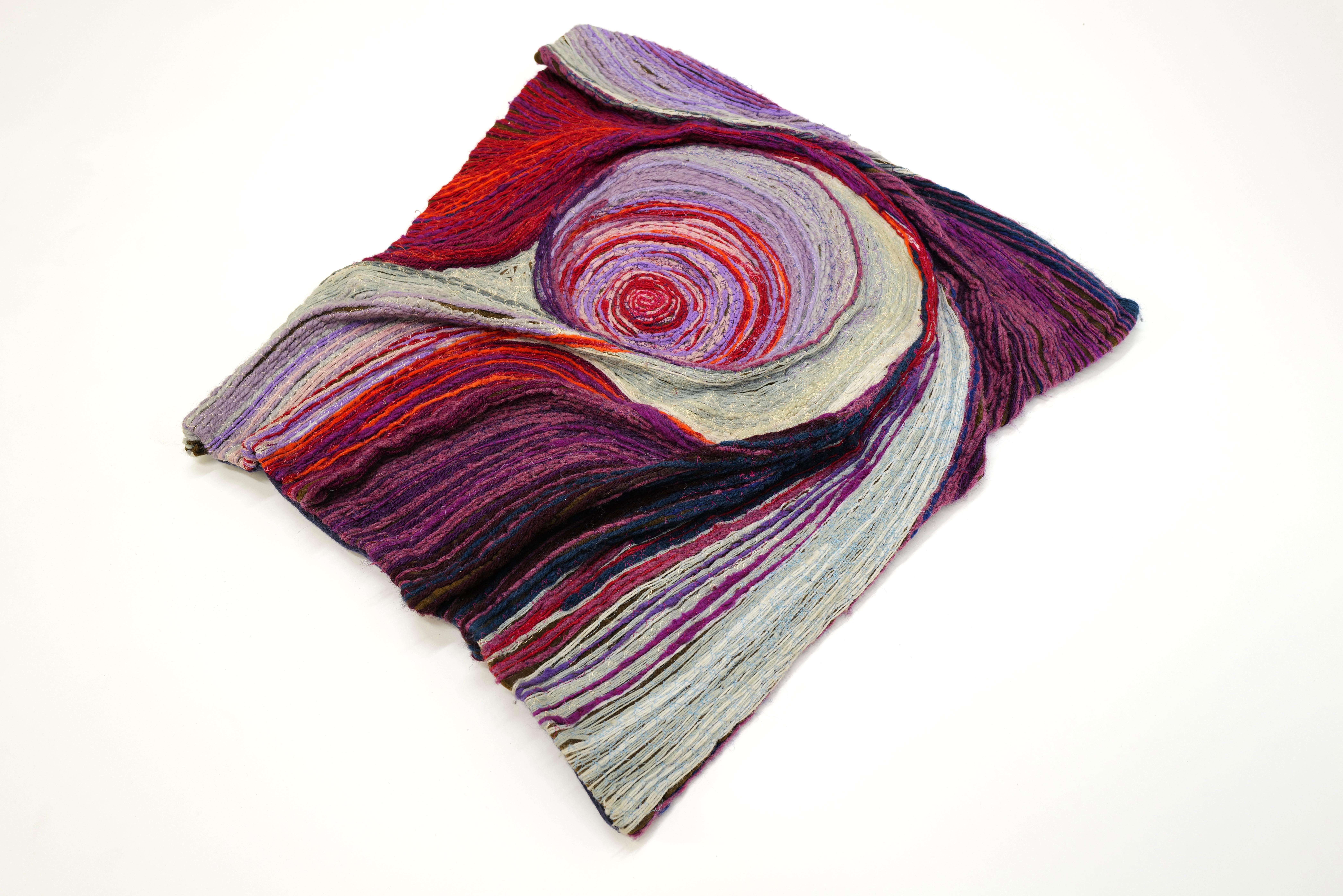 Janet Kuemmerlein's stunning fiber art piece, measuring 24”x 26”, is a captivating exploration of color and texture. With a mesmerizing palette ranging from vibrant purples to fiery reds, interspersed with hints of white or cream, the artwork evokes