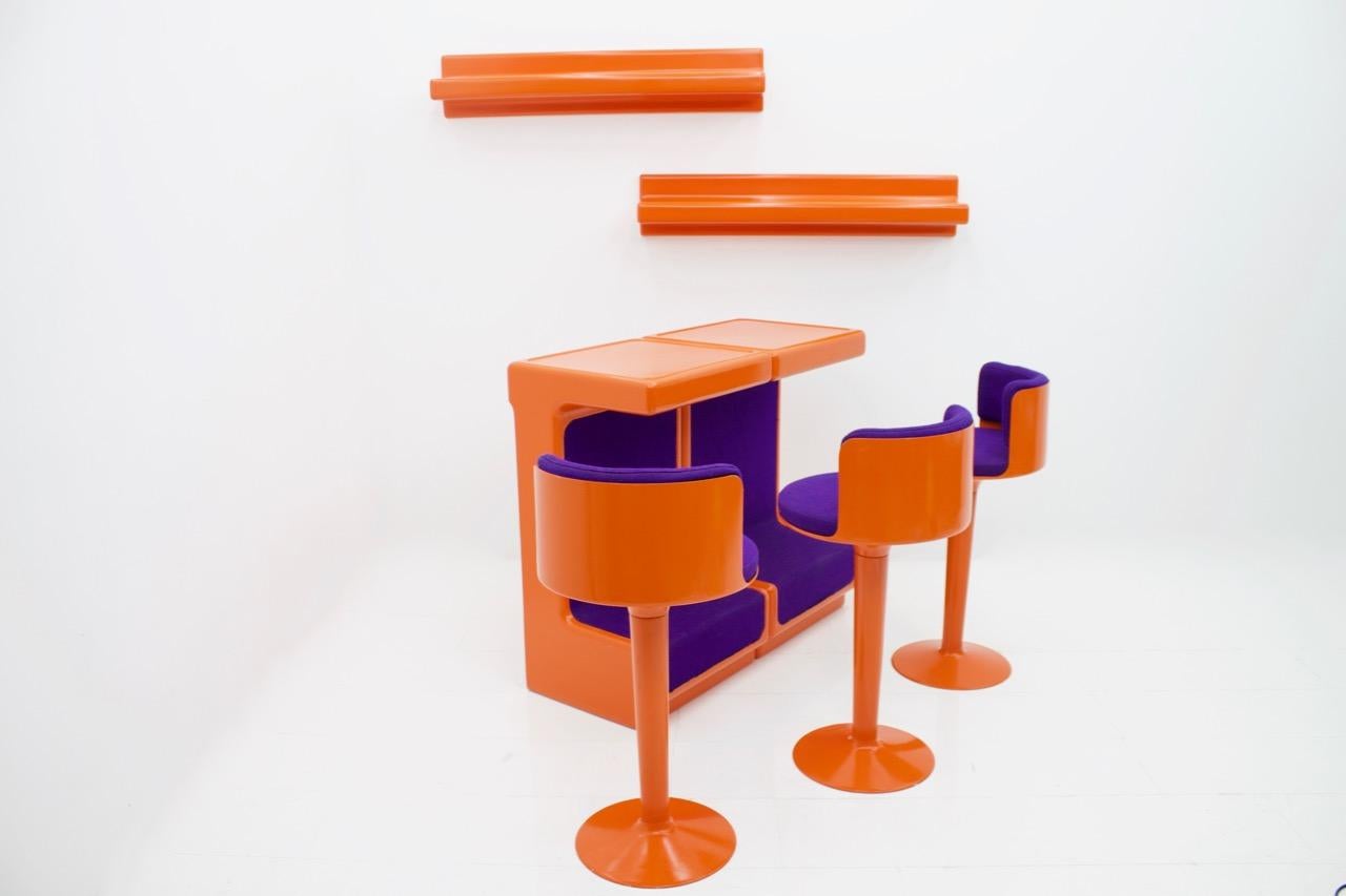 Bar-Set von Wolfgang Feierbach bestehend aus 2 modularen Theken:: 2 Wandregalen und 3 drehbaren Barhockern. 
Die Möbel sind aus Fiberglas gefertigt:: orange lackiert und teilweise mit lilafarbenem Stoff bespannt. 
Der Zustand ist gut mit