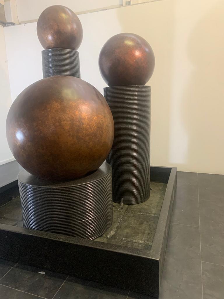A splendid and imposing work by Ravi Sing consisting of a metal and fibreglass basin, here are three cylinders with bodies decorated with relief lines of different sizes and heights. Above the cylinders are copper-coated fibreglass spheres. For