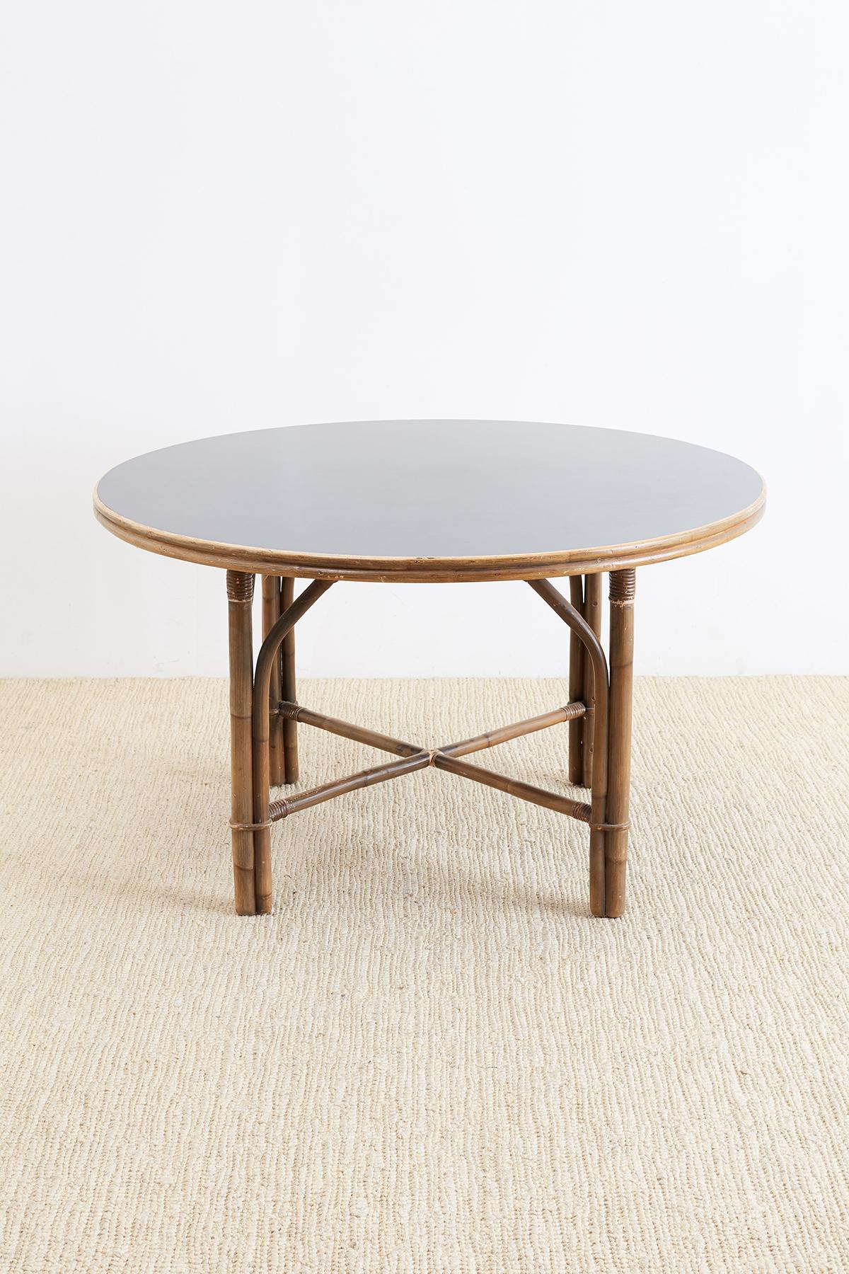 Mid-Century Modern period rattan and formica dining table or patio table by Ficks Reed. Features a large round formica black top with a double bamboo edge. The table is supported by round bamboo legs conjoined with stretchers and reinforced with