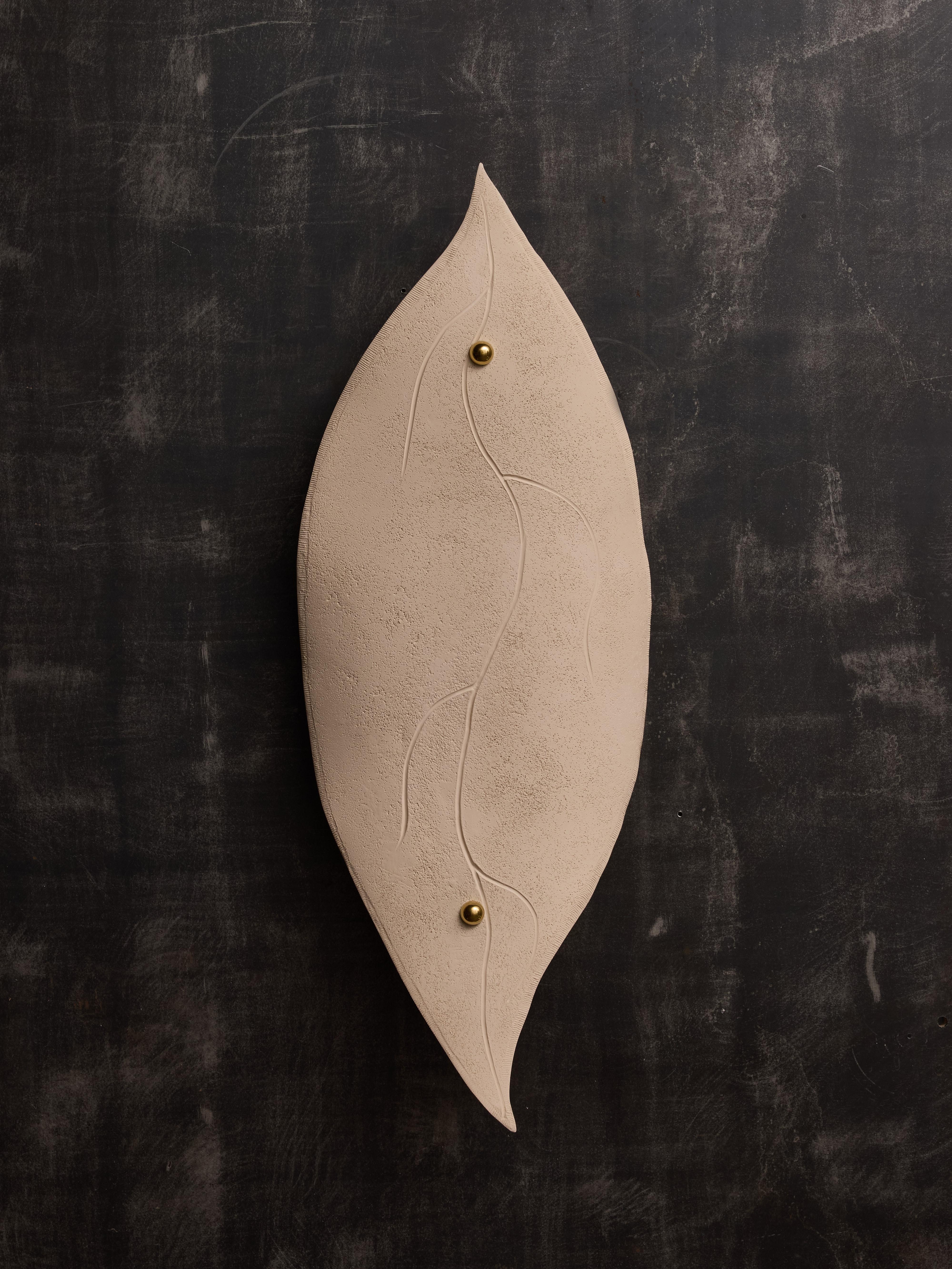 Los apliques de pared creados por la artista francesa Françoise Bergaglia presentan una placa trasera de latón que soporta dos fuentes de luz, elegantemente adornada con un panel decorativo de loza con forma de hoja de ficus.