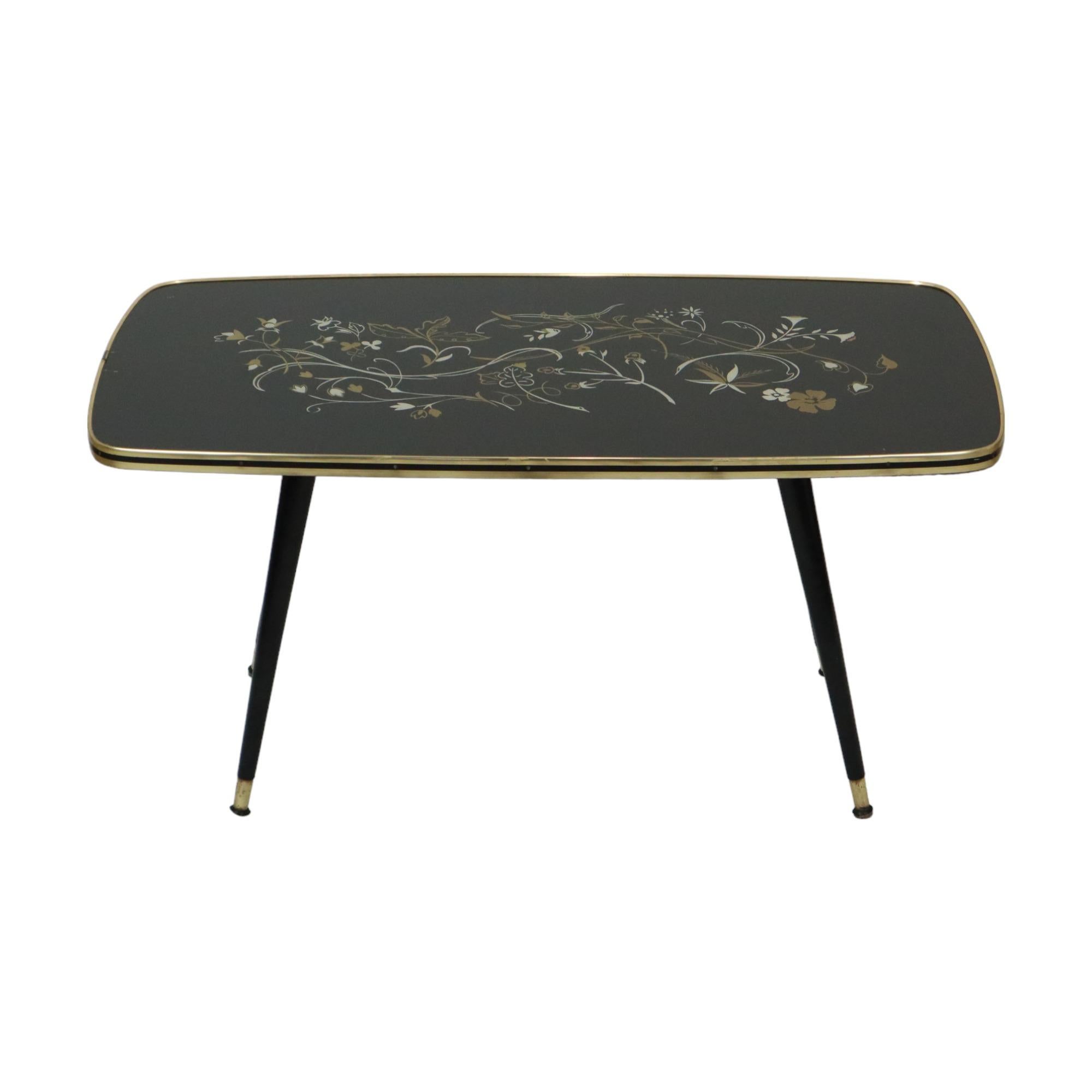 Table basse vintage avec plateau en verre, plateau noir décoré d'un motif floral et d'une bande dorée tout autour, pieds coniques finis en cuivre, design des années 50, Belgique.

Le style de cette table est également appelé 