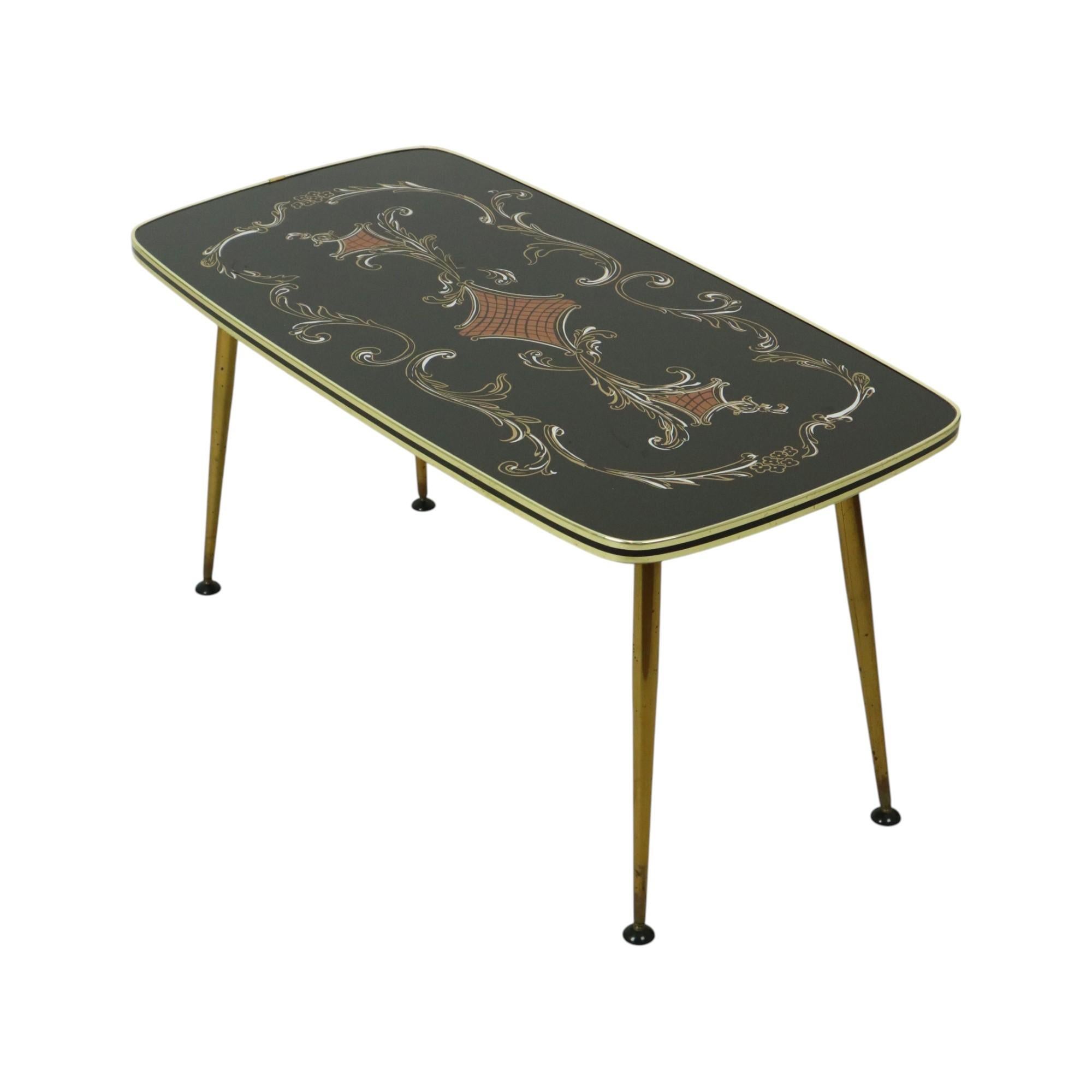 Mesa de centro vintage con tablero de cristal, tablero negro decorado con un motivo floral de acanto y una banda dorada alrededor con una línea negra central, patas cónicas chapadas en cobre con pies ajustables inclinables en el extremo, diseño de