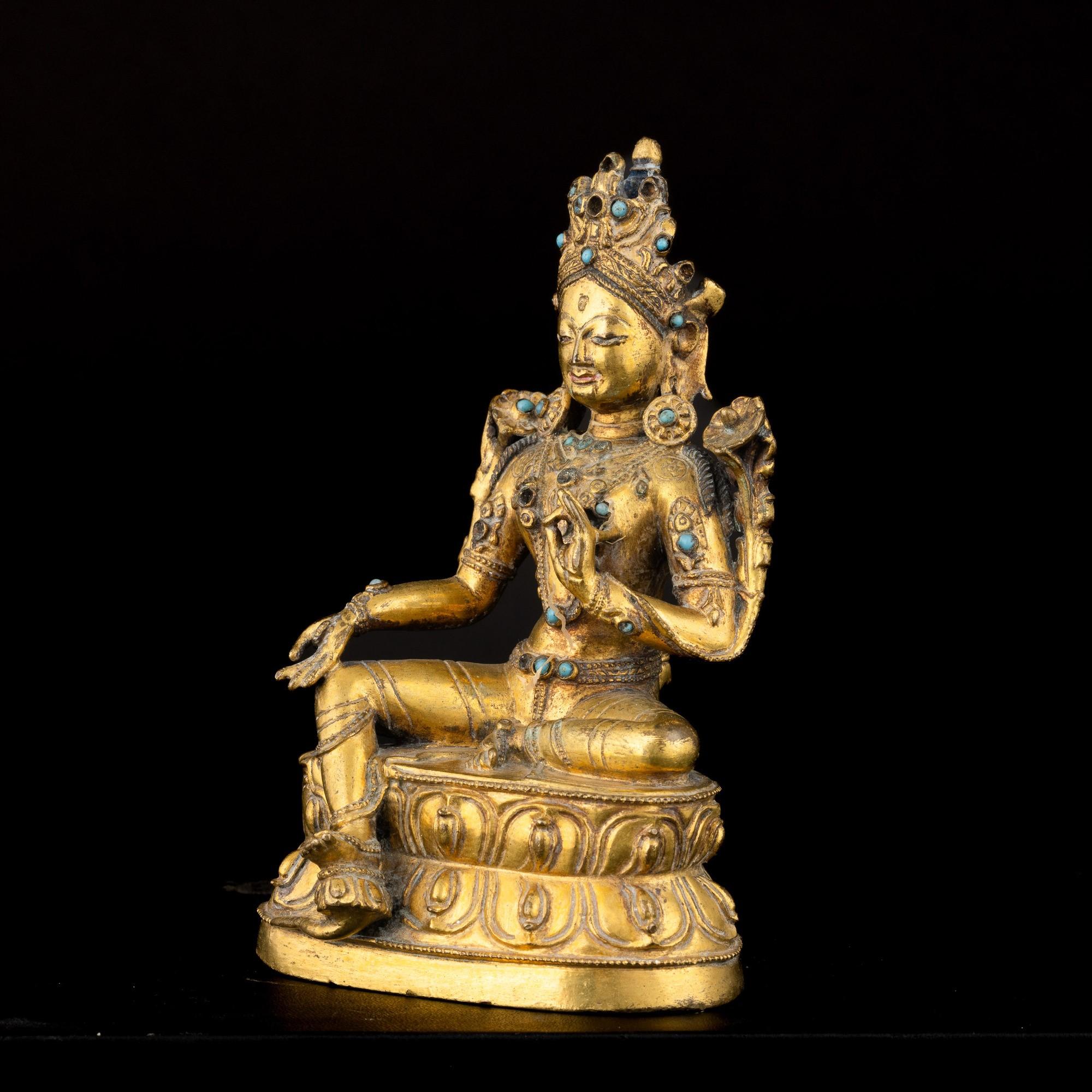 Esta figura de Tara, de bronce dorado, data de los siglos XVII-XVIII y procede del Tíbet. La deidad aparece sentada sobre una flor de loto doble, símbolo de pureza espiritual. La escultura presenta restos de policromía e injertos de turquesa, que
