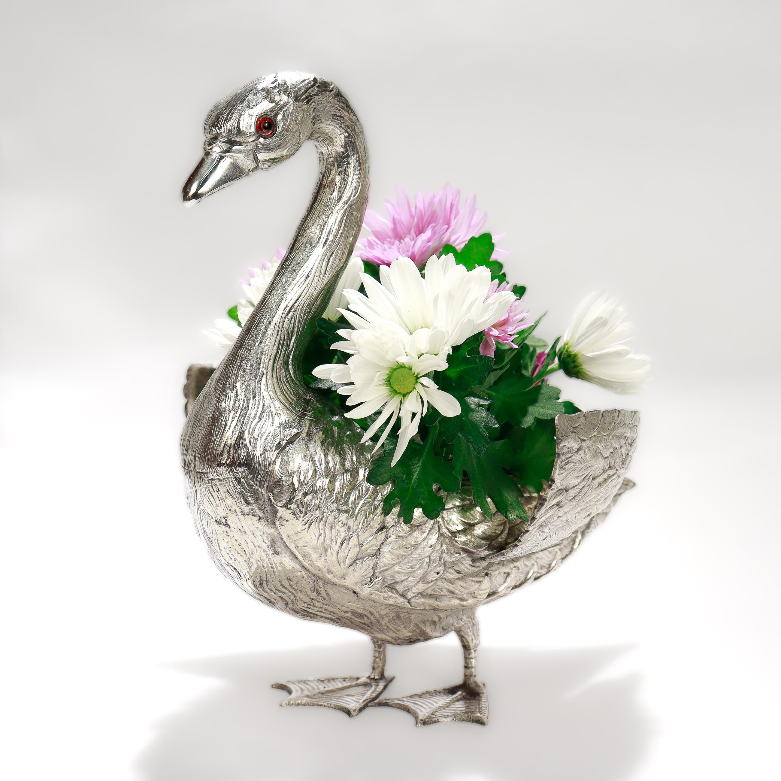 Ein feines figurales Mittelstück.

Aus Alpaka 'Silber'.

In Form eines stehenden Schwans mit Glasaugen und klappbaren Flügeln. 

Die Flügel lassen sich öffnen und geben den Blick auf einen abnehmbaren Blumenfrosch oder -halter frei. 

Markiert