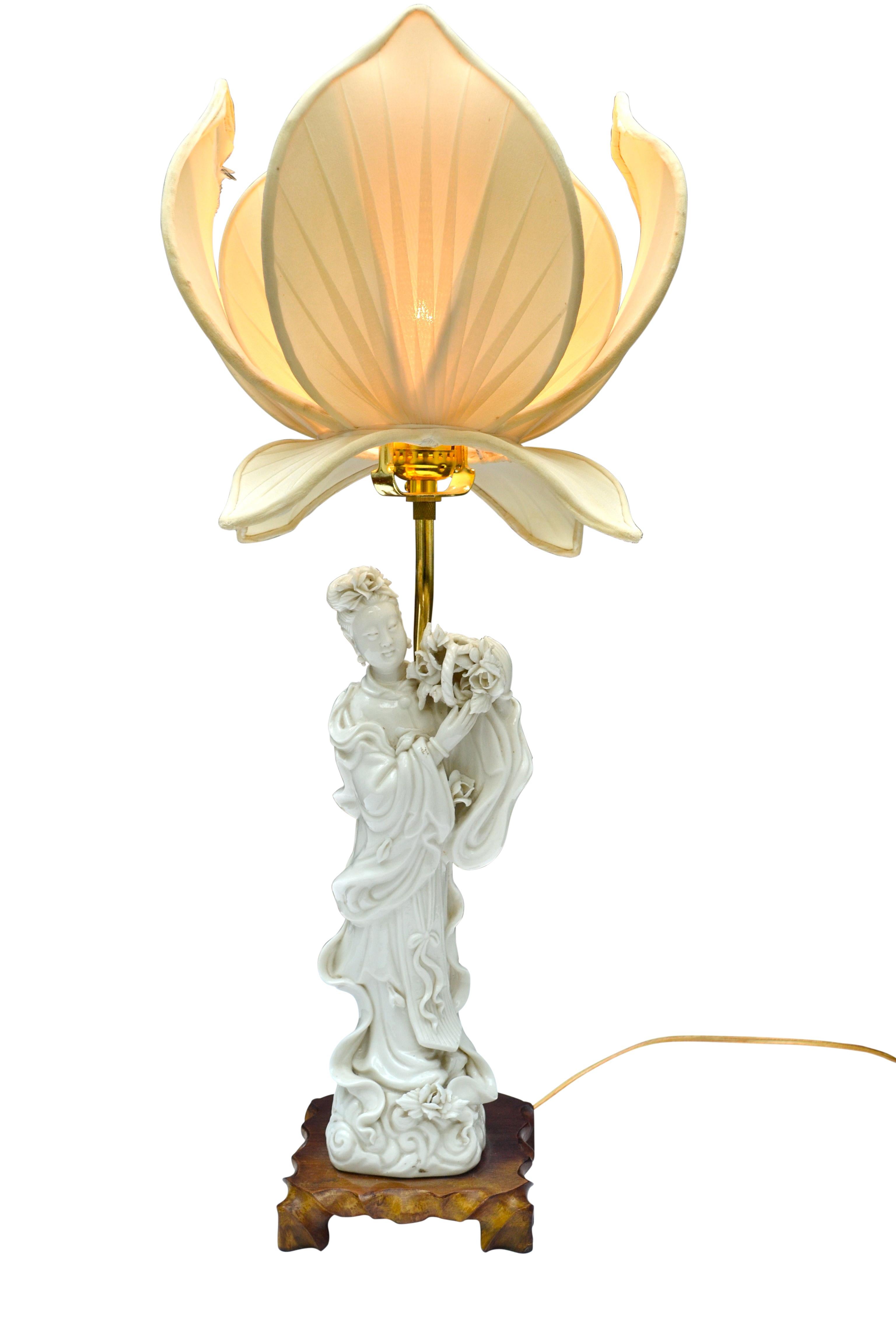 A Blanc De Chine lamp depicting a standing figure Quan Yin holding aloft a basket of roses set on a wooden base and featuring a unique original silk shade in the shape of a lotus flower.
  
