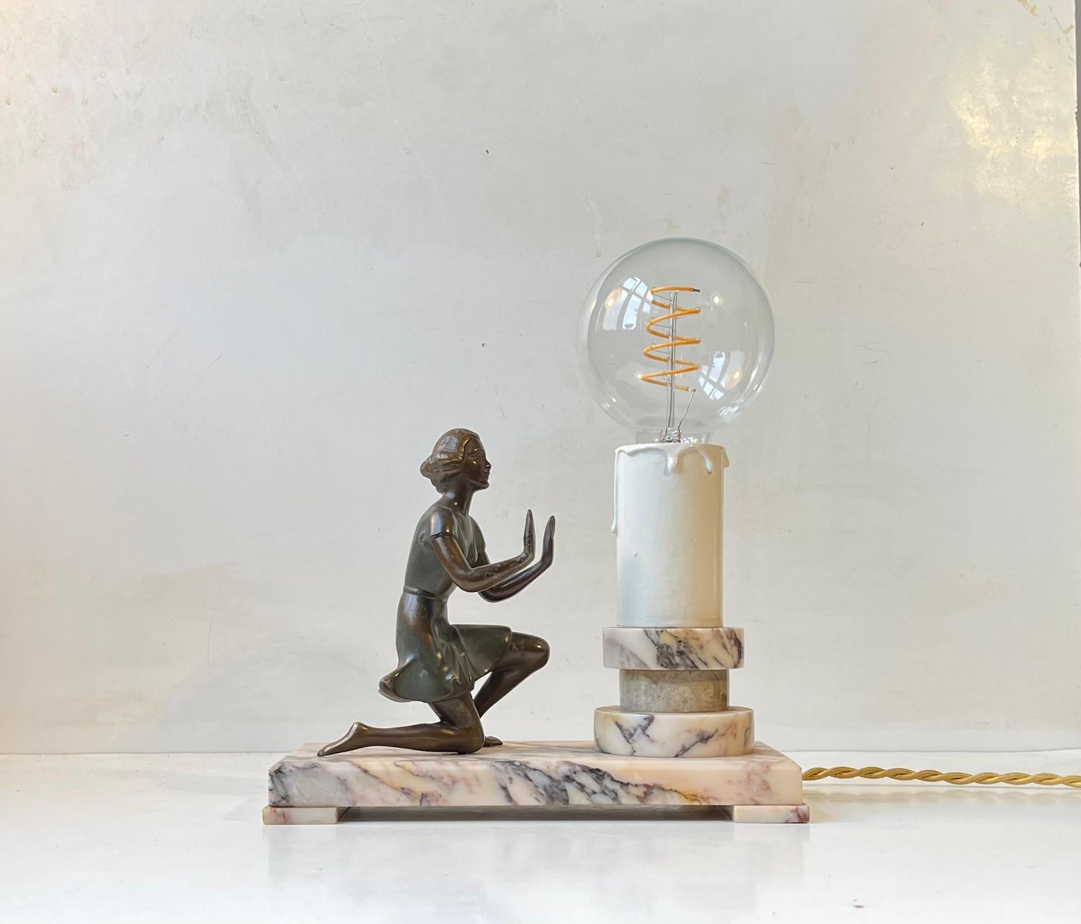 Lampe de table ornée d'une figurine, avec une base architecturale en marbre onyx, une fausse bougie stylisée et une sculpture/figurine en bronze qui se réchauffe les mains devant la 
