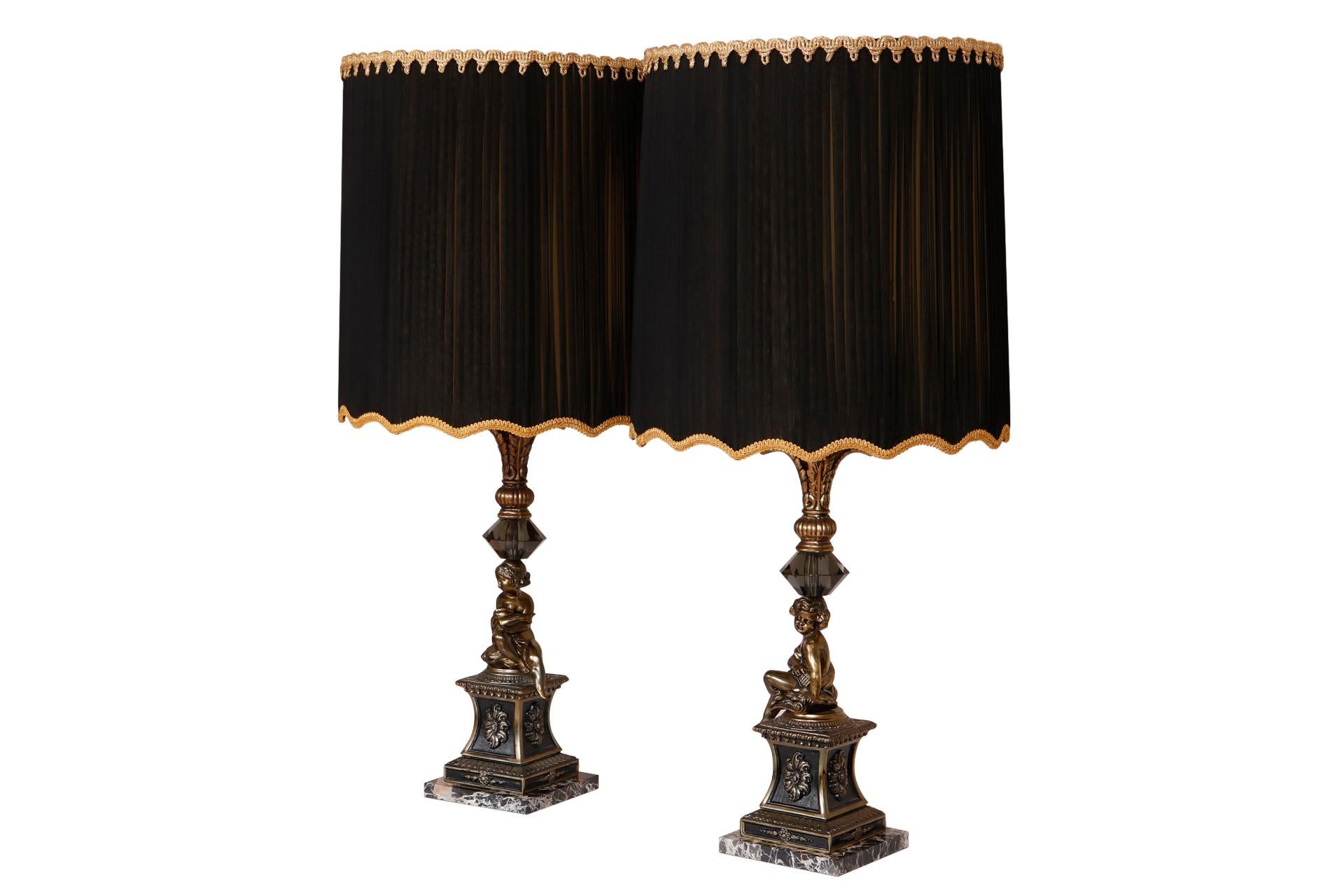 A pair of figural French Empire style table lamps. Rouched black shades are trimmed with a rope twist detail and gold gimp. Columns are topped with cast brass acanthus plumes above large cut glass crystals. Opposing cast cherubs sit on painted metal
