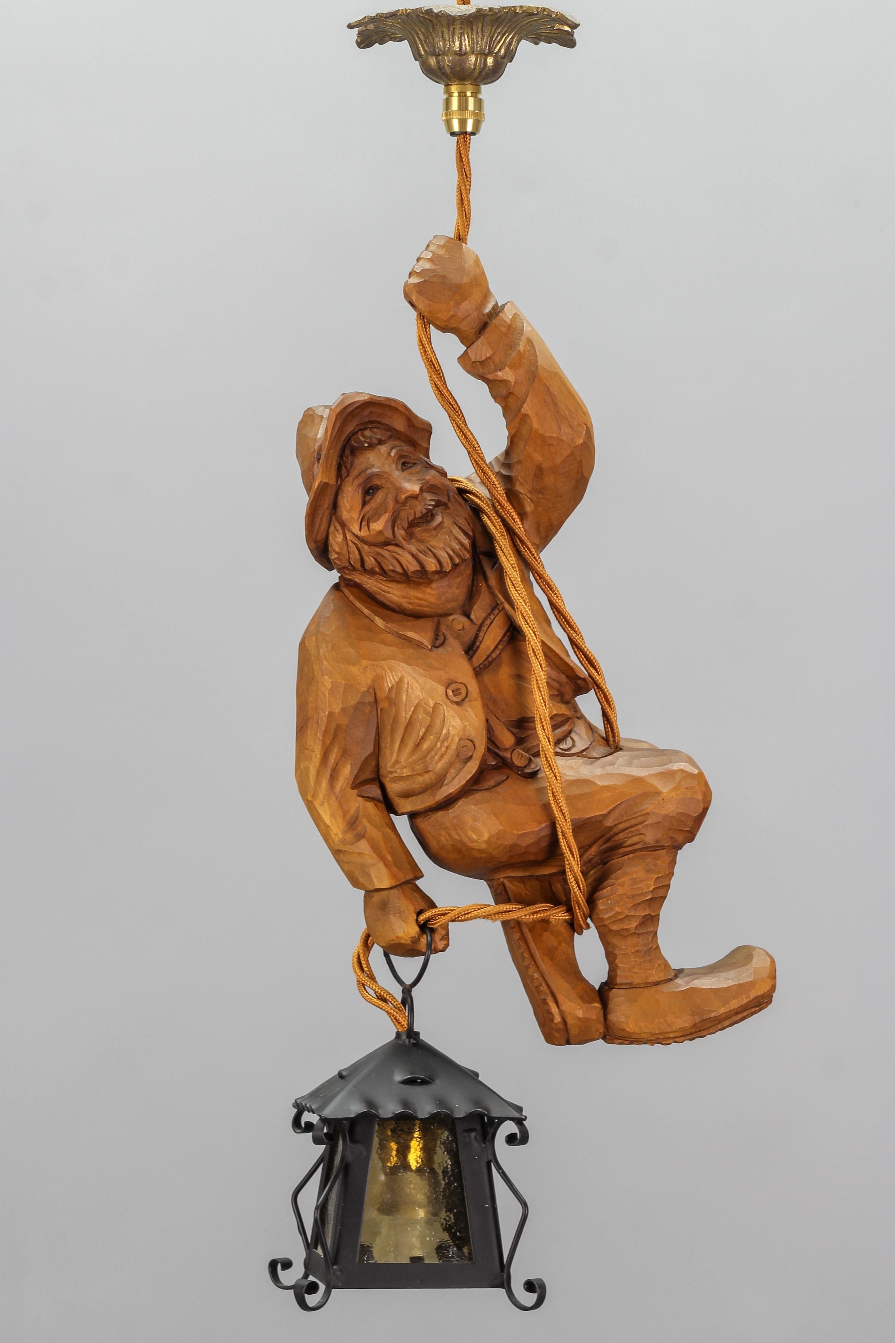 Cette ravissante lampe suspendue figurative allemande présente une figure en bois de tilleul sculptée à la main représentant un alpiniste souriant et chaleureux. L'alpiniste en bois sculpté se tient à une corde et tient une lanterne dans une main.