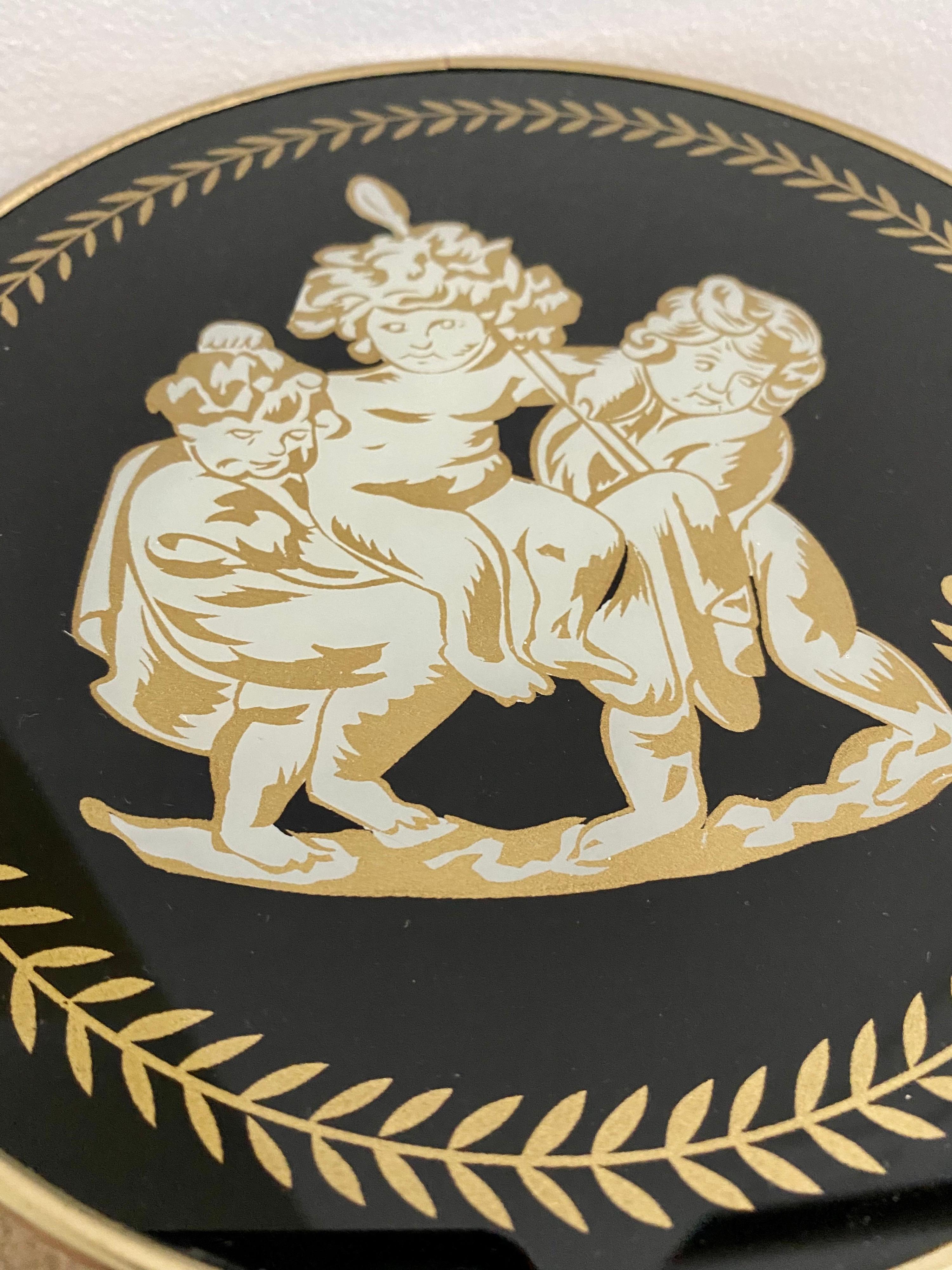 Set of four reverse painted glass putti cherub laurel leaf wall art plaques. These figural round art pieces are framed in gold brass frames and feature black glass with gilt painted accents. 