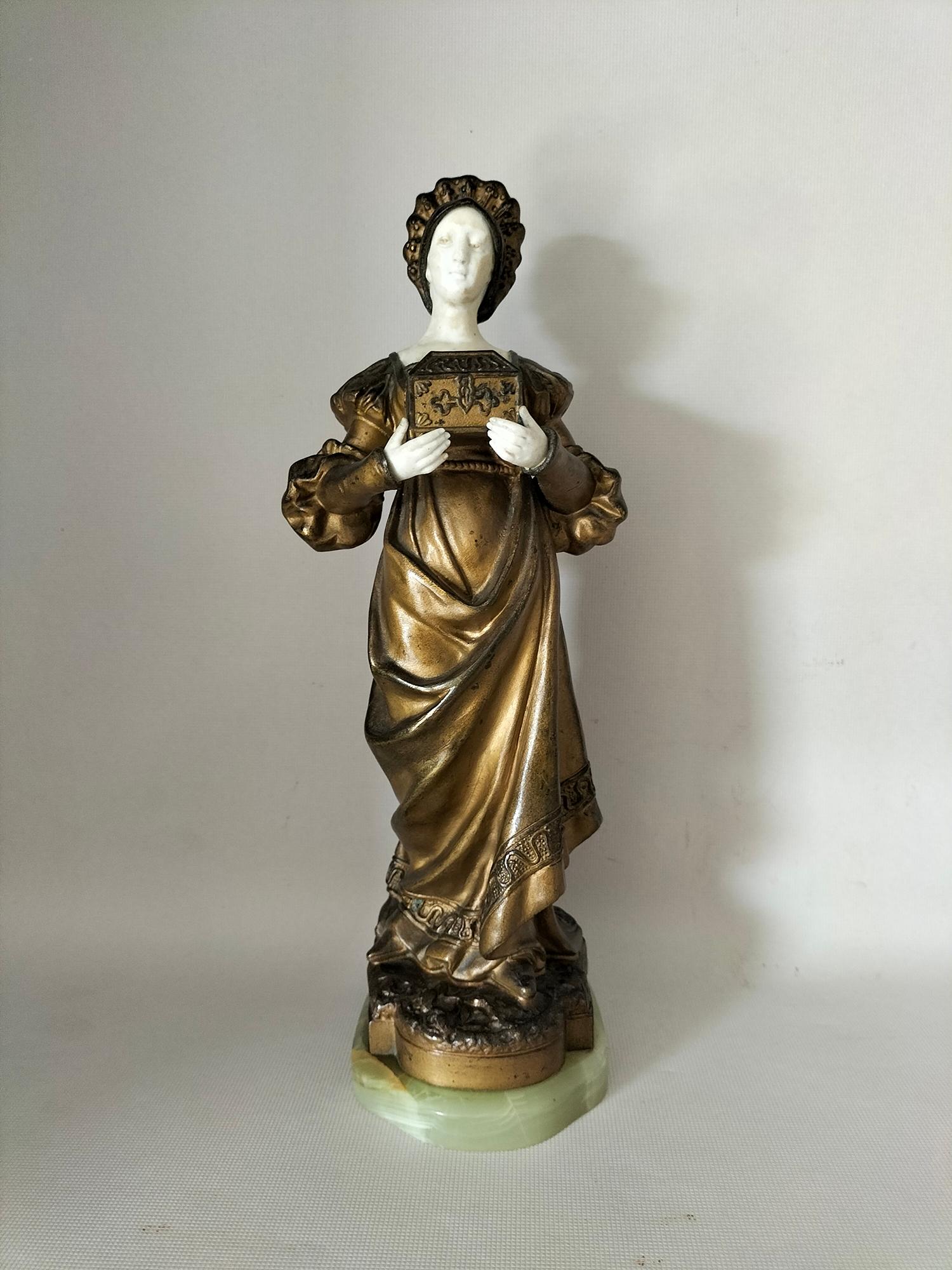 Calamine figure patinated in various golden tones, representing a woman in a period dress, carrying a chest.
Her hands and face are biscuit, with some wear to the face.
The base is in jade
Lady with a chest ( probably Marguerite of Faust. 
Faust is