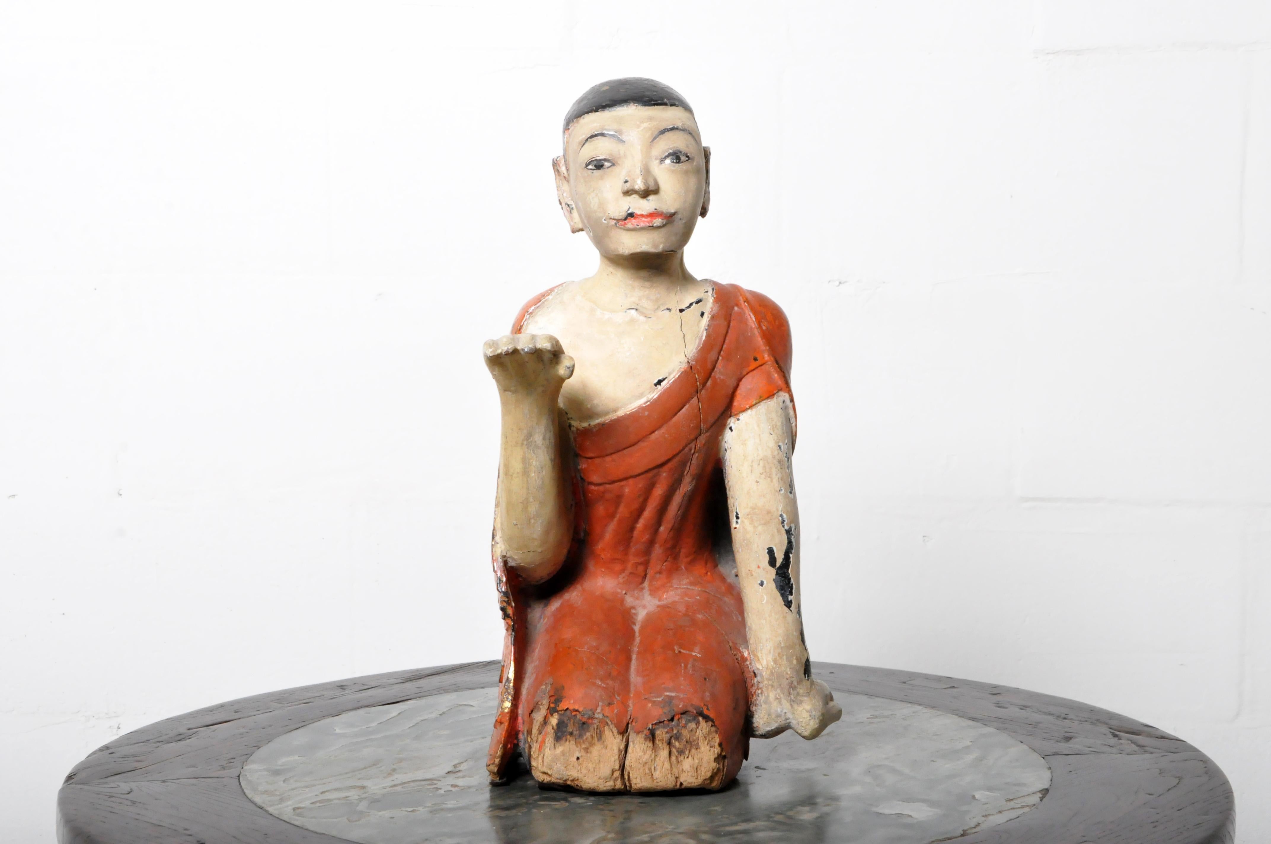 A teak wood figure of a young monk in a posture of devotion and with a facial expression is one of piety and innocence. The piece is covered in natural red cinnabar lacquer and polychrome. There is some damage to the wood from moisture; this is