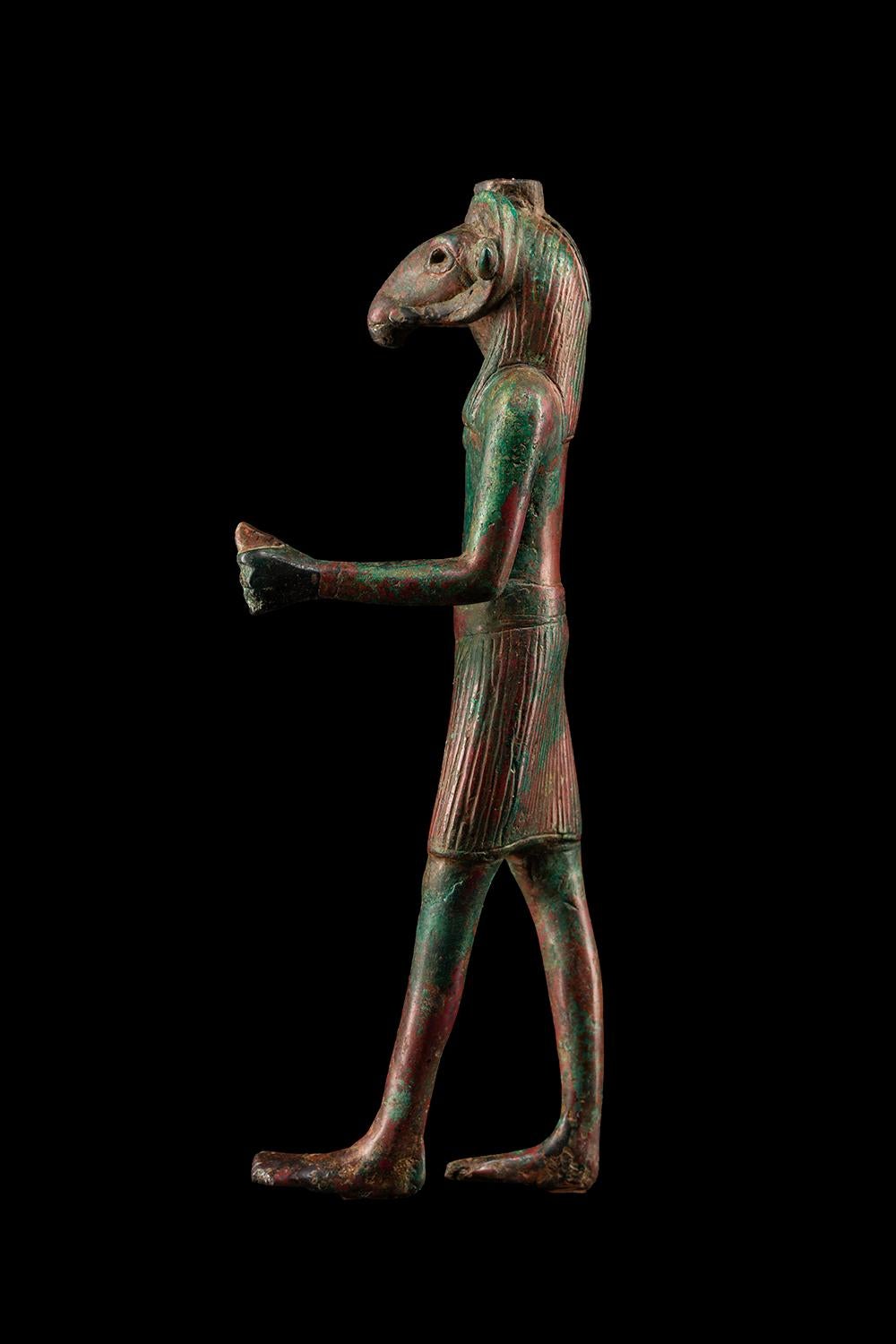 Figura del dio egizio Khnum che cammina, in bronzo. Il dio è raffigurato come un uomo con una testa di ariete, con due grandi corna che si curvano verso l'interno e in avanti verso il viso, nello stile delle successive rappresentazioni di Khnum (le