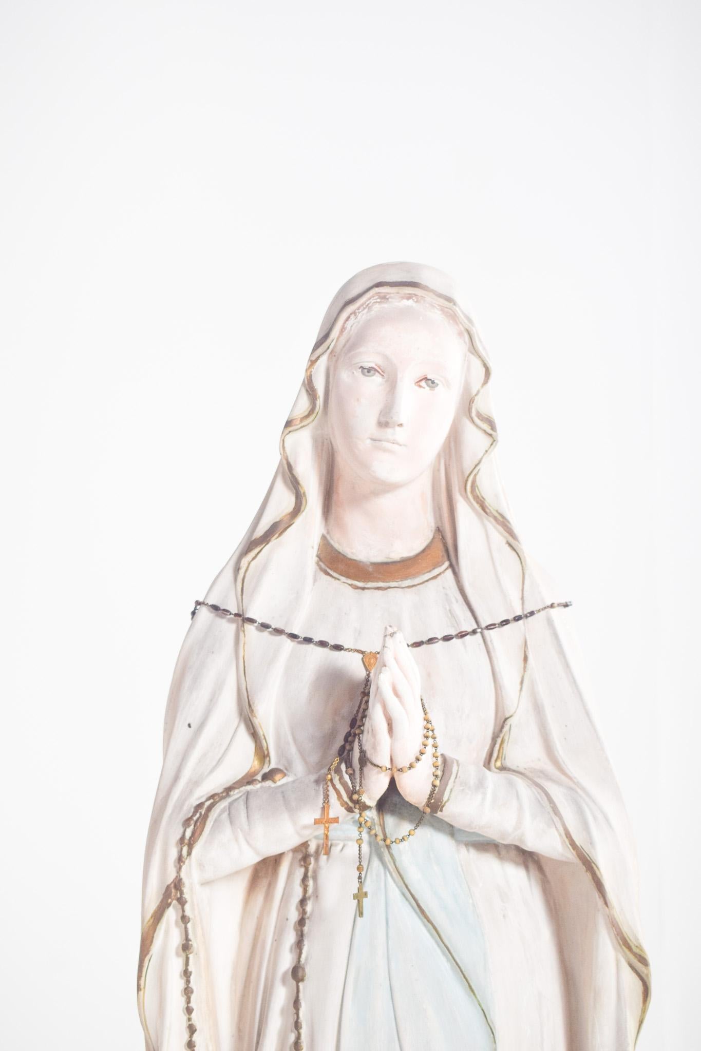Figura di Nostra Signora di Lourdes, in ceramica. L'immagine raffigura la Vergine Maria in lunghe vesti nei toni del bianco e dell'azzurro, con dettagli dorati sui bordi. Le sue mani sono unite in preghiera e tengono un rosario con grani e una croce