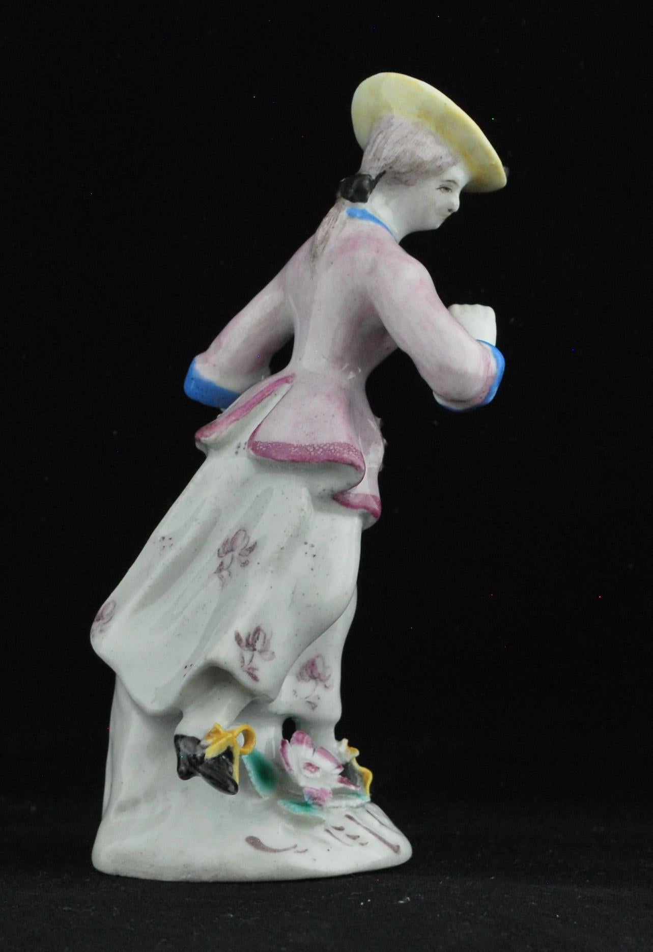Petite figure d'une jeune femme portant une jupe blanche tachetée de puce, légèrement soulevée de la main gauche, une veste rose pâle avec col et poignets bleus opaques typiques de l'arc.

Nous l'avons appelée 