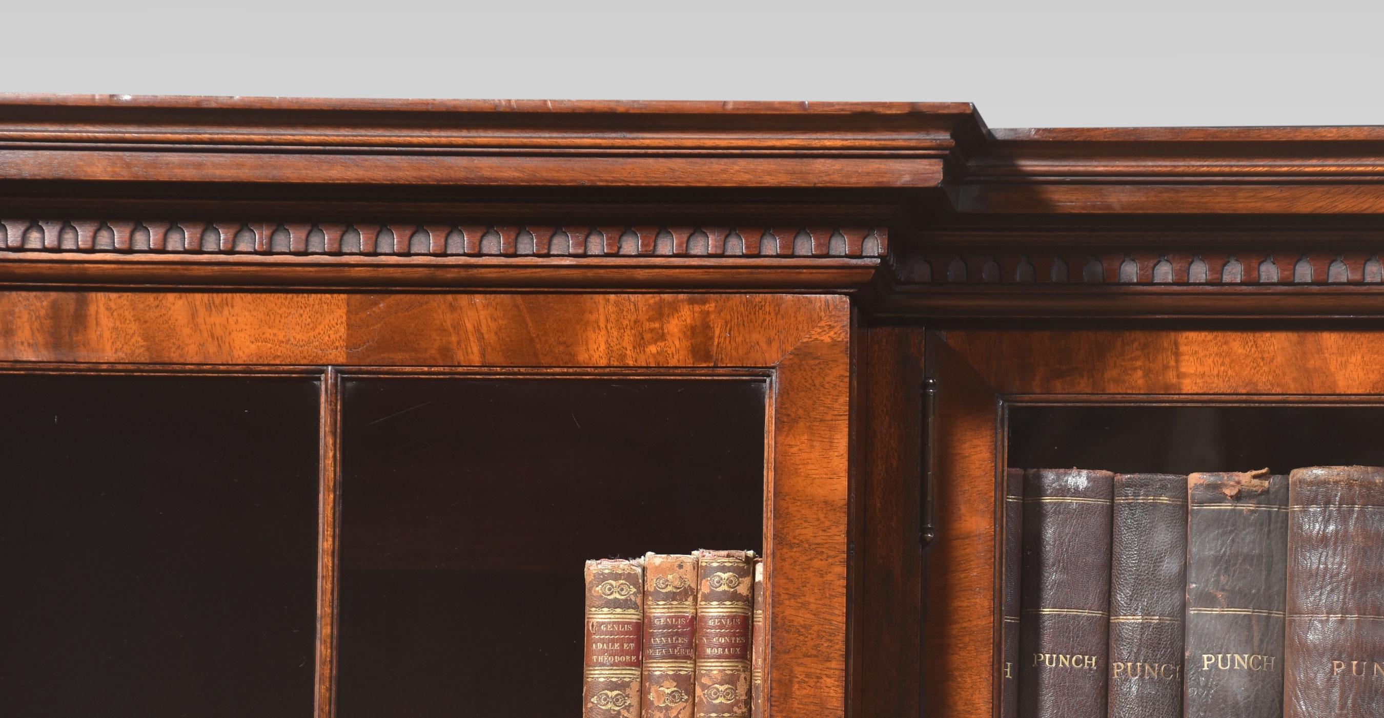 Figured mahogany inverted breakfront bookcase, the projecting moulded cornice above two large central glazed doors opening to reveal an adjustable shelved interior. Flanked by two further large glazed doors with oval inlaid panels. Opening to reveal
